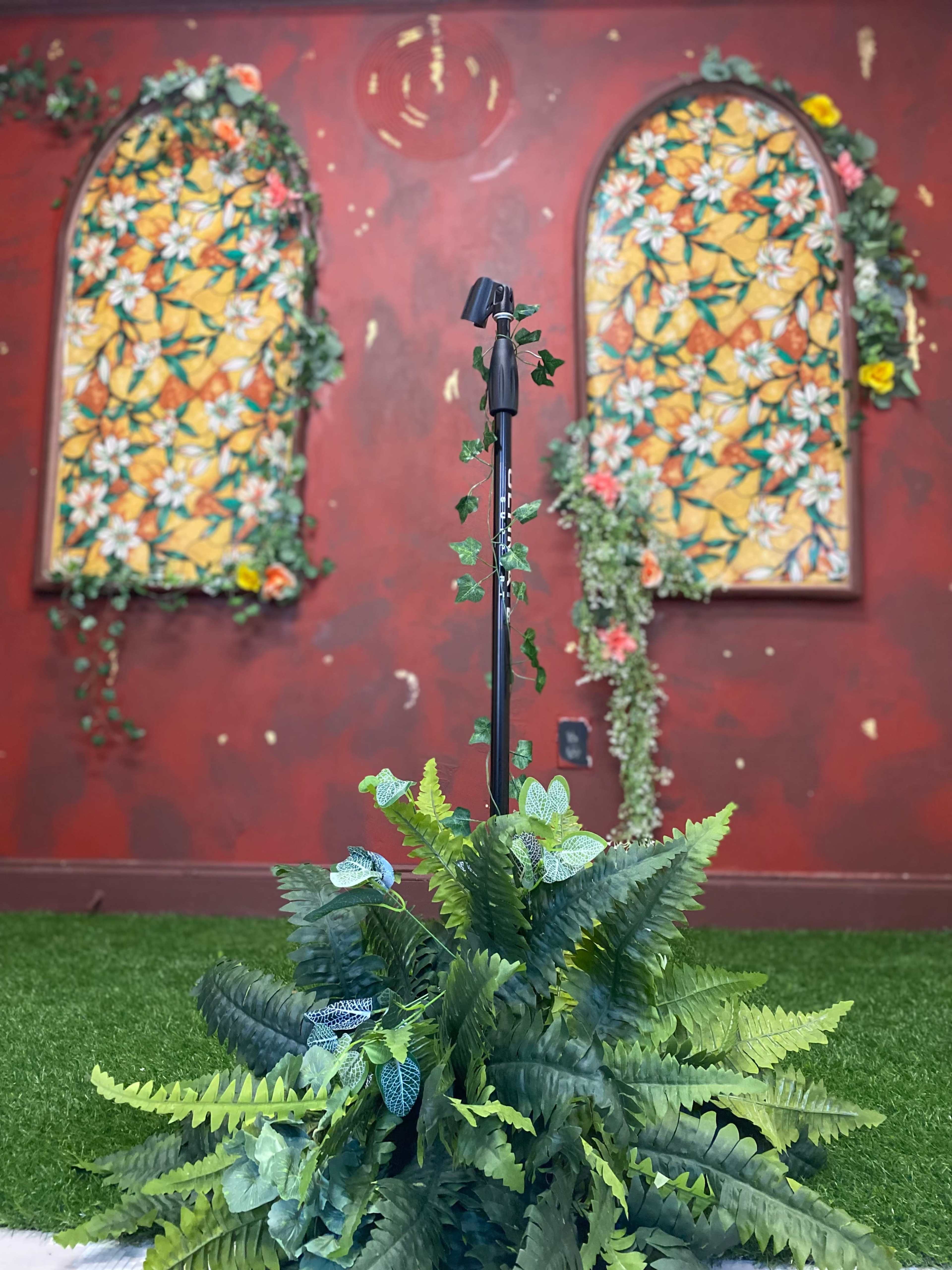 A microphone stand is positioned in front of two flower-adorned windows on a red wall, with greenery placed around a faux grass floor.