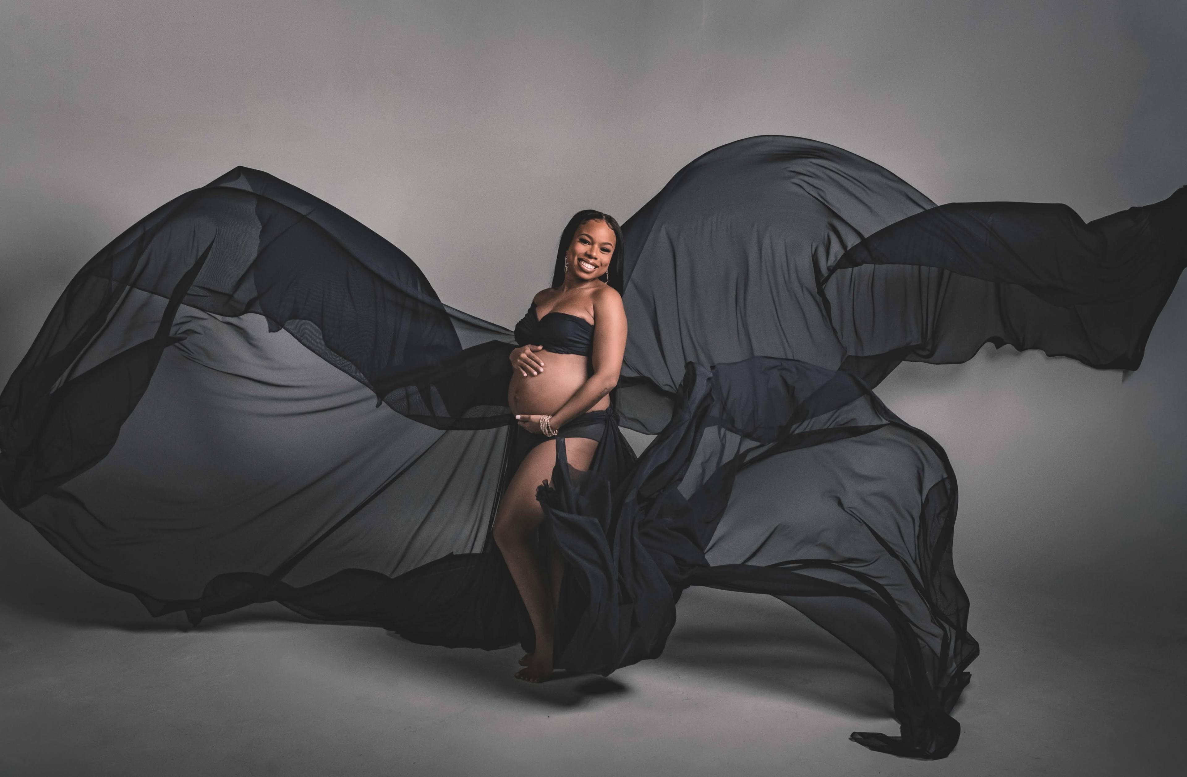 A pregnant woman stands gracefully, holding her belly while draped in flowing black fabric in a studio setting.
