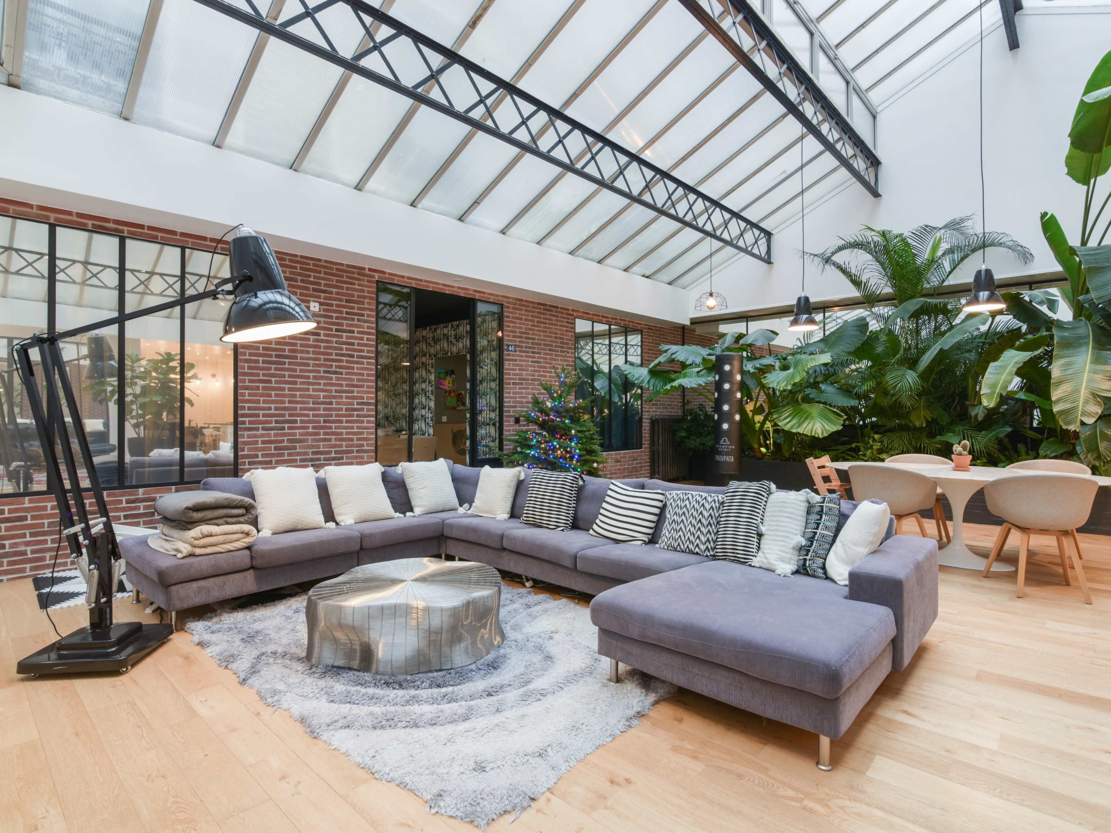 The image shows a spacious modern living area featuring a large sectional sofa, a round coffee table, and abundant greenery, with a glass ceiling allowing natural light to enter.
