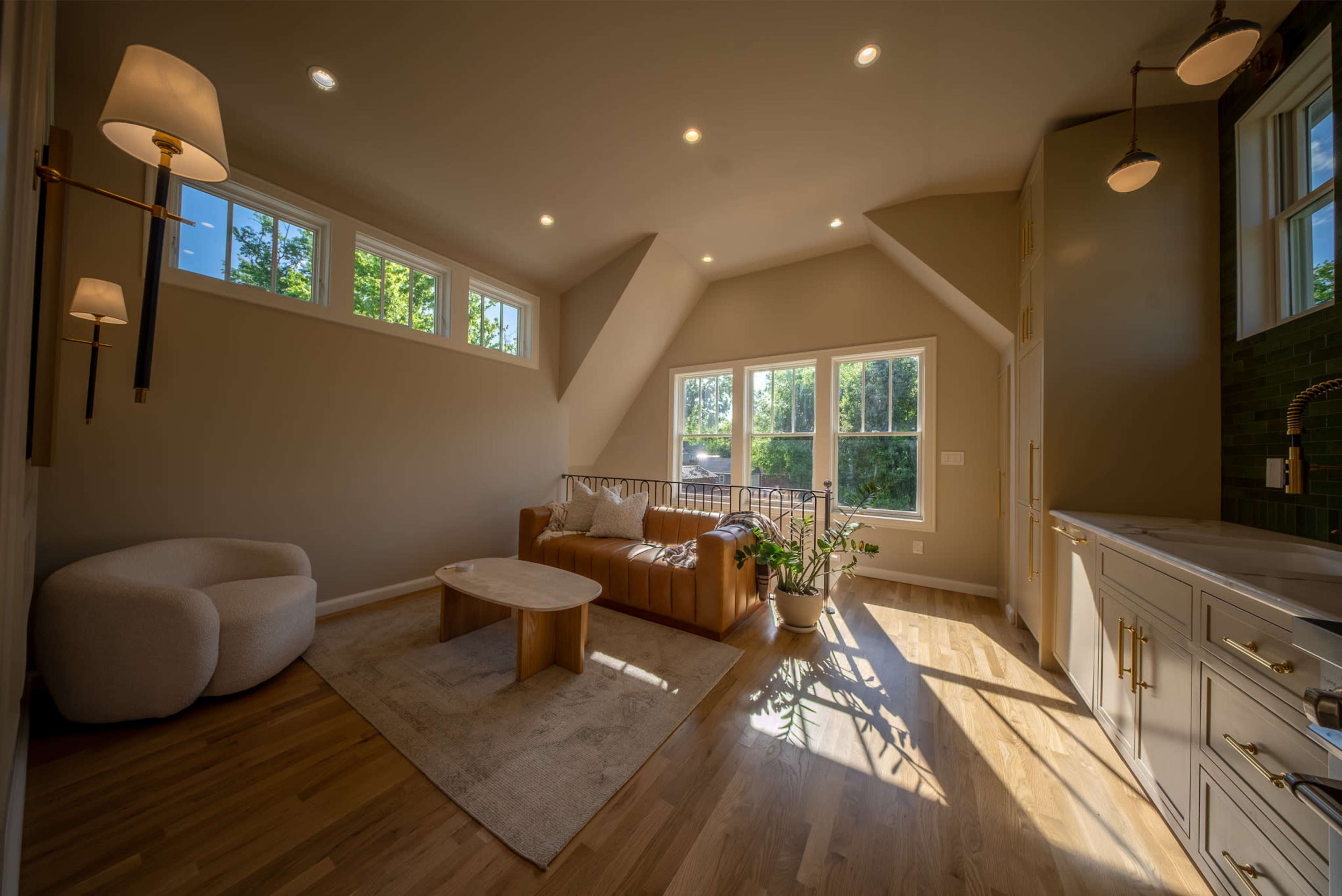 The image shows a modern living room with a light-colored wood floor, a leather sofa, a round coffee table, and large windows that allow natural light to fill the space.