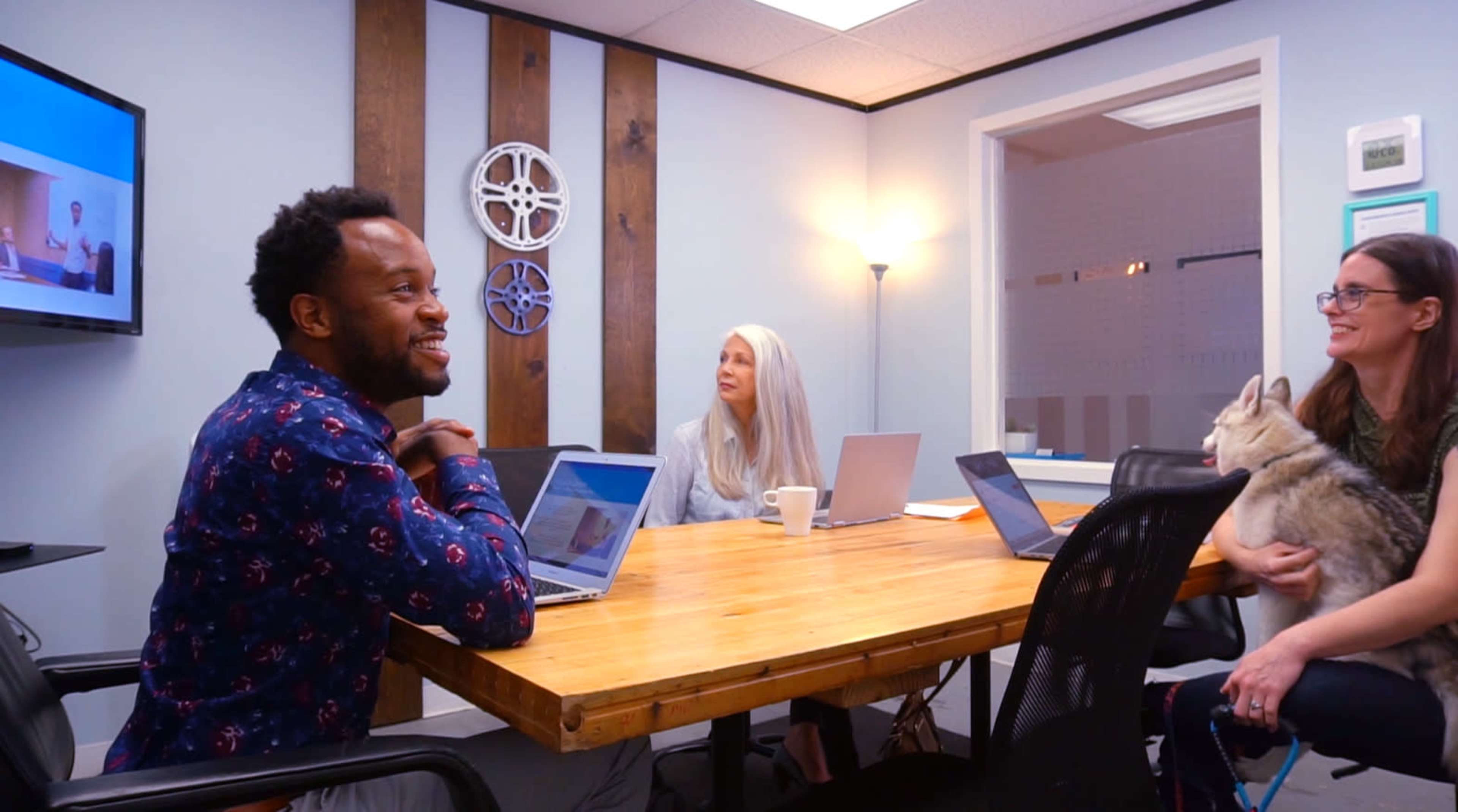 A group of three people sits around a wooden table in a meeting room, working on laptops and engaging in conversation, while a dog rests in one person's arms.