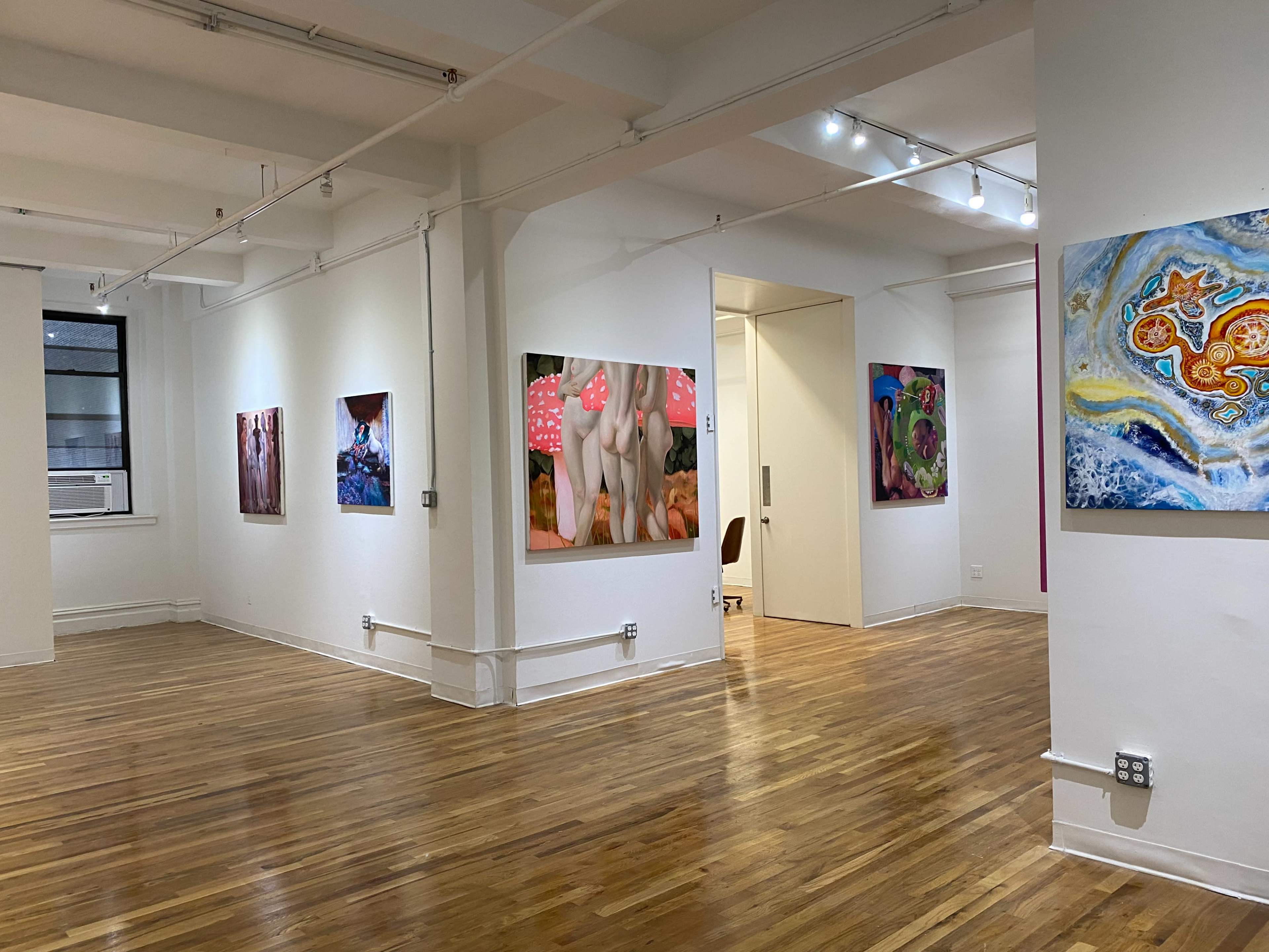 A spacious art gallery with several colorful paintings displayed on the white walls and polished wooden flooring.