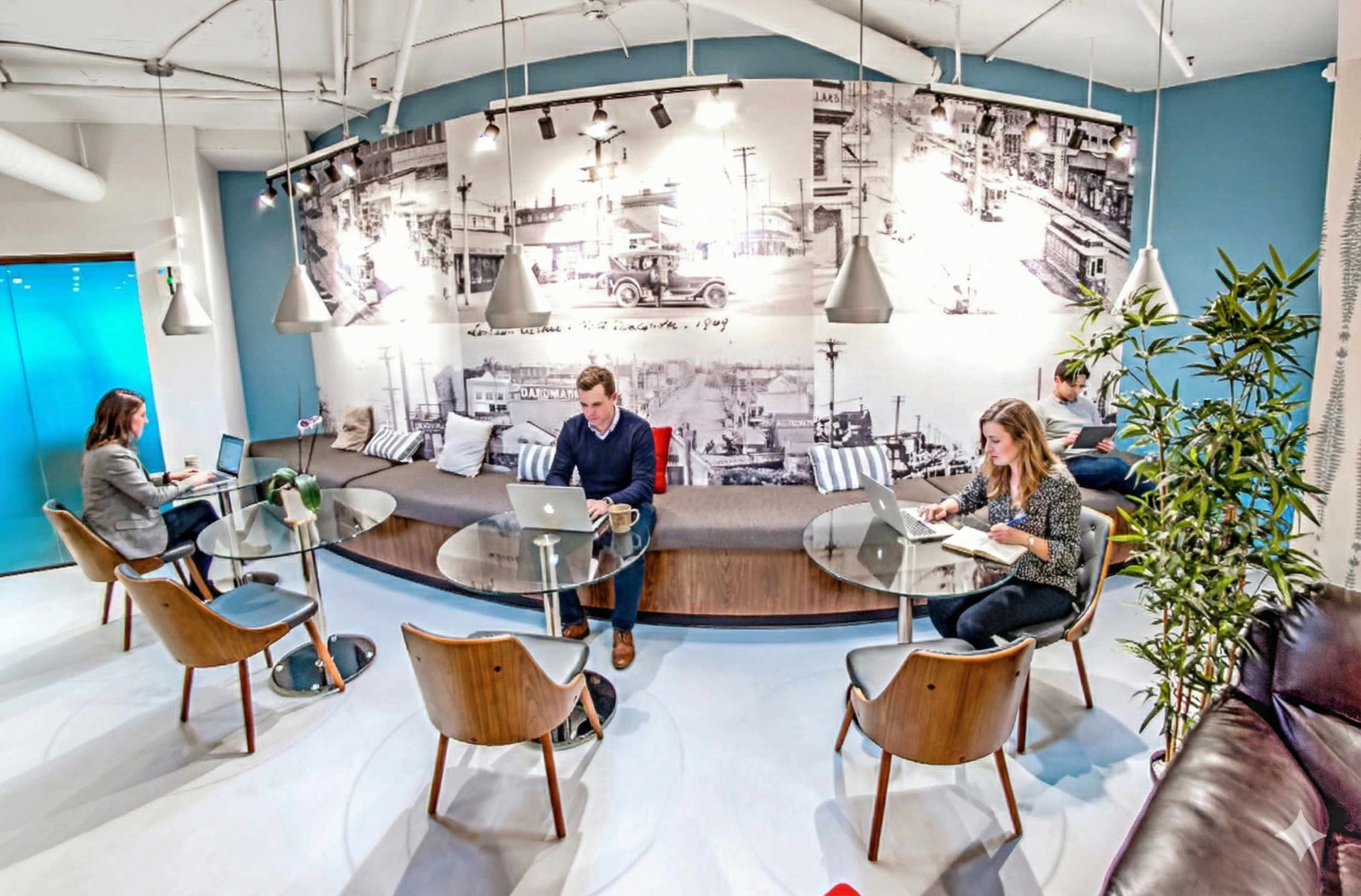 A modern coworking space features several people working on laptops at stylish tables surrounded by a historical mural.