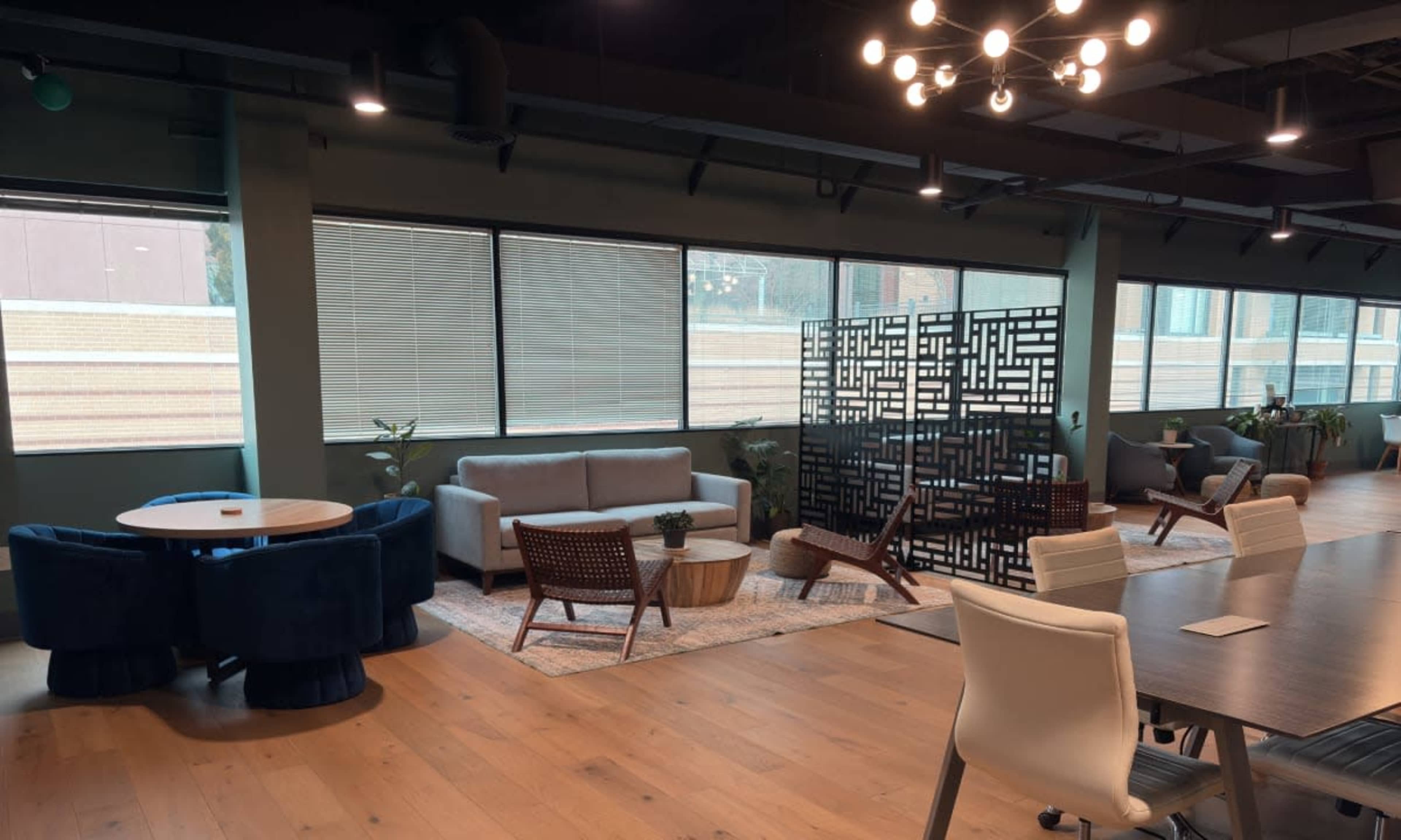 The image shows a modern office space featuring a seating area with a gray sofa, wooden chairs, a table, and a decorative partition, all illuminated by natural light from large windows.