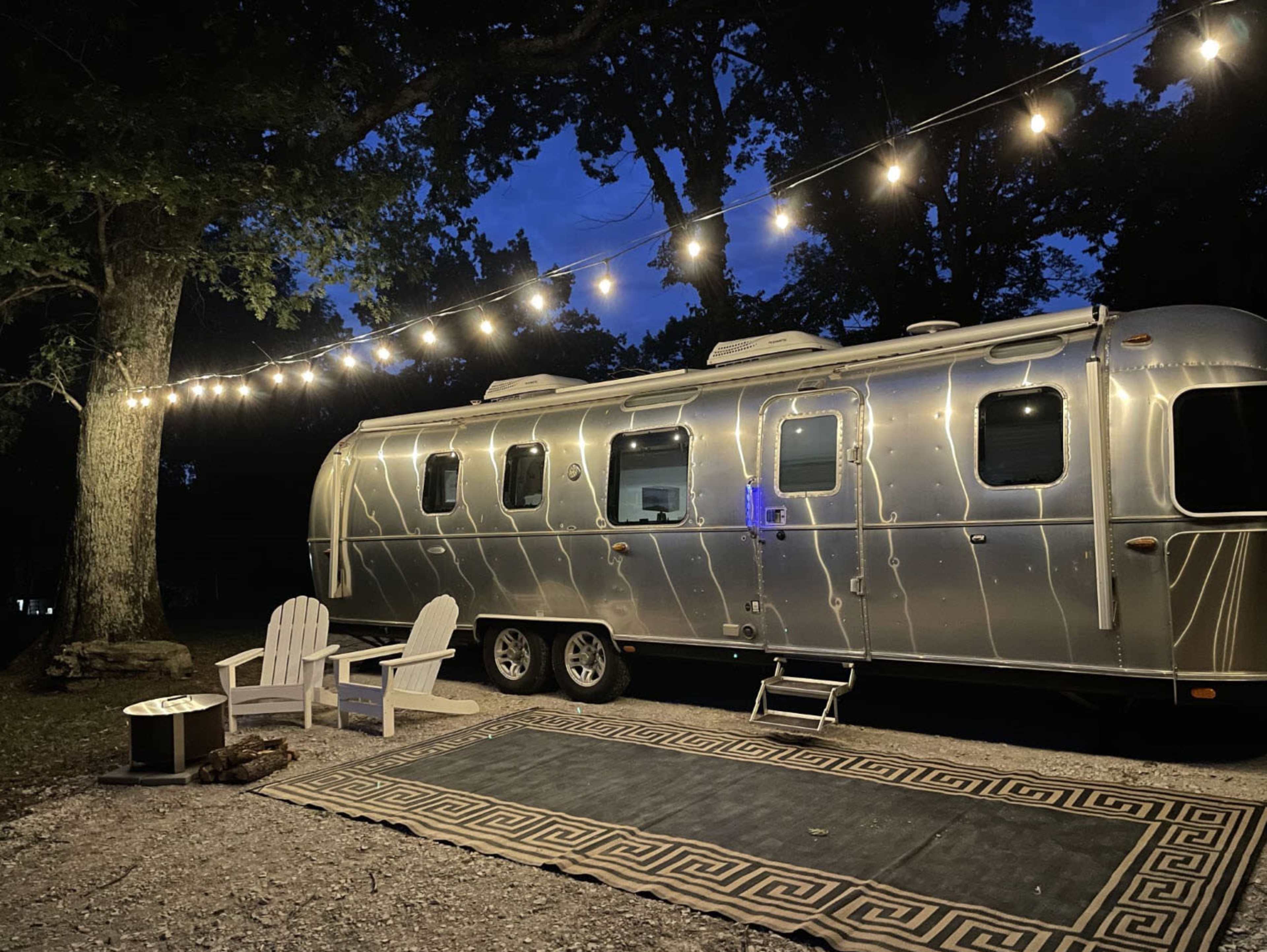 A polished silver Airstream trailer is parked under string lights next to a fire pit and two Adirondack chairs on a patterned rug.