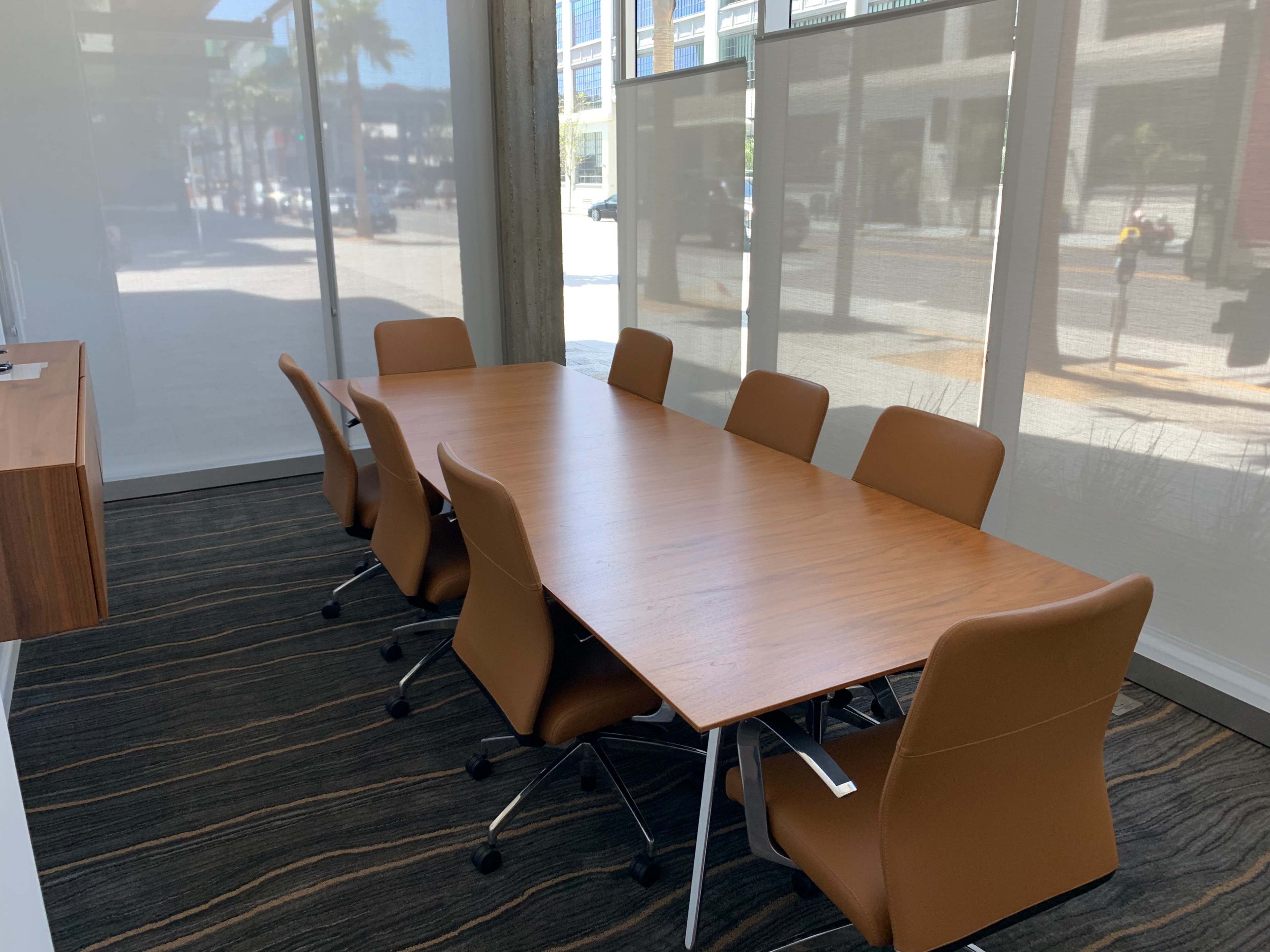 A conference room features a long wooden table surrounded by eight brown chairs, with large windows allowing natural light to enter.