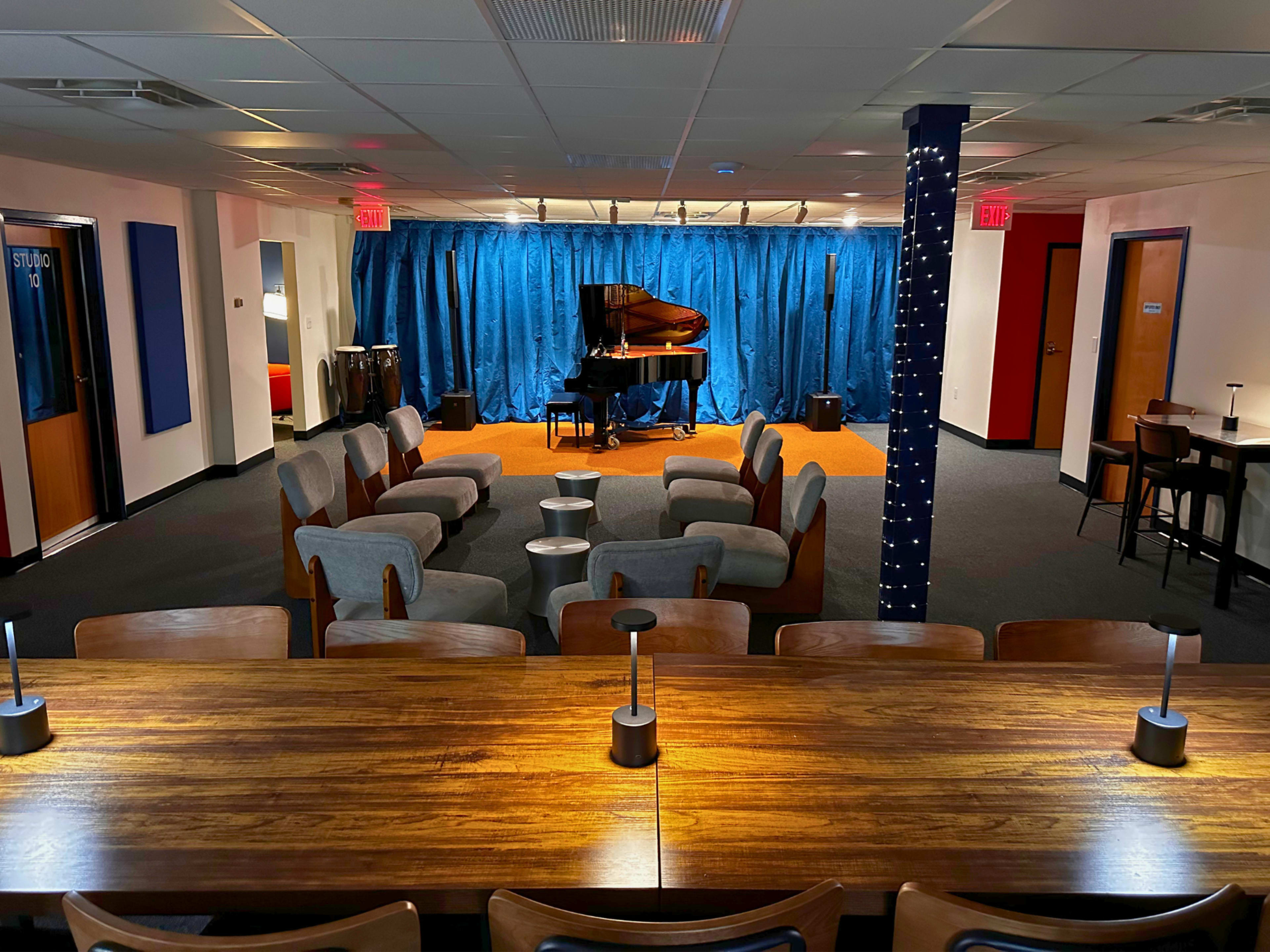 The image shows a spacious room with a grand piano on stage, surrounded by chairs and a long wooden table with modern light fixtures.