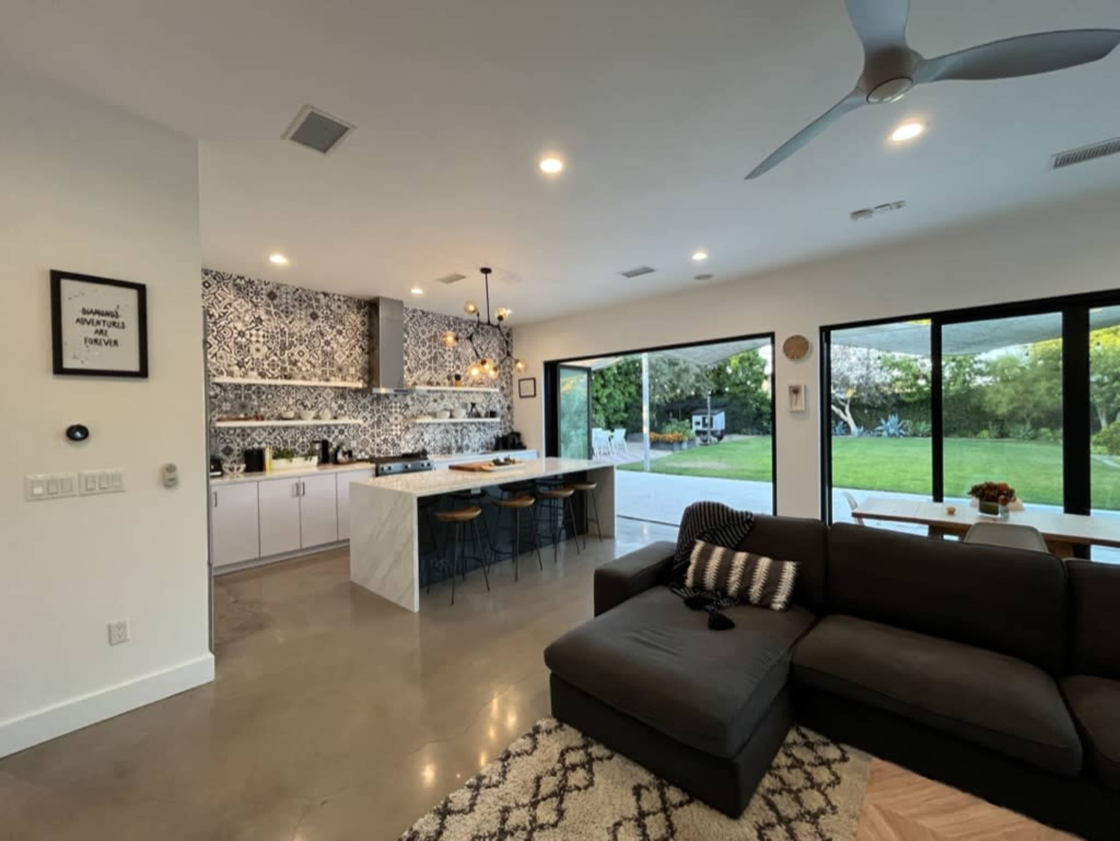 A modern kitchen and living area with a large sectional sofa, a patterned backsplash, and large windows overlooking a green outdoor space.