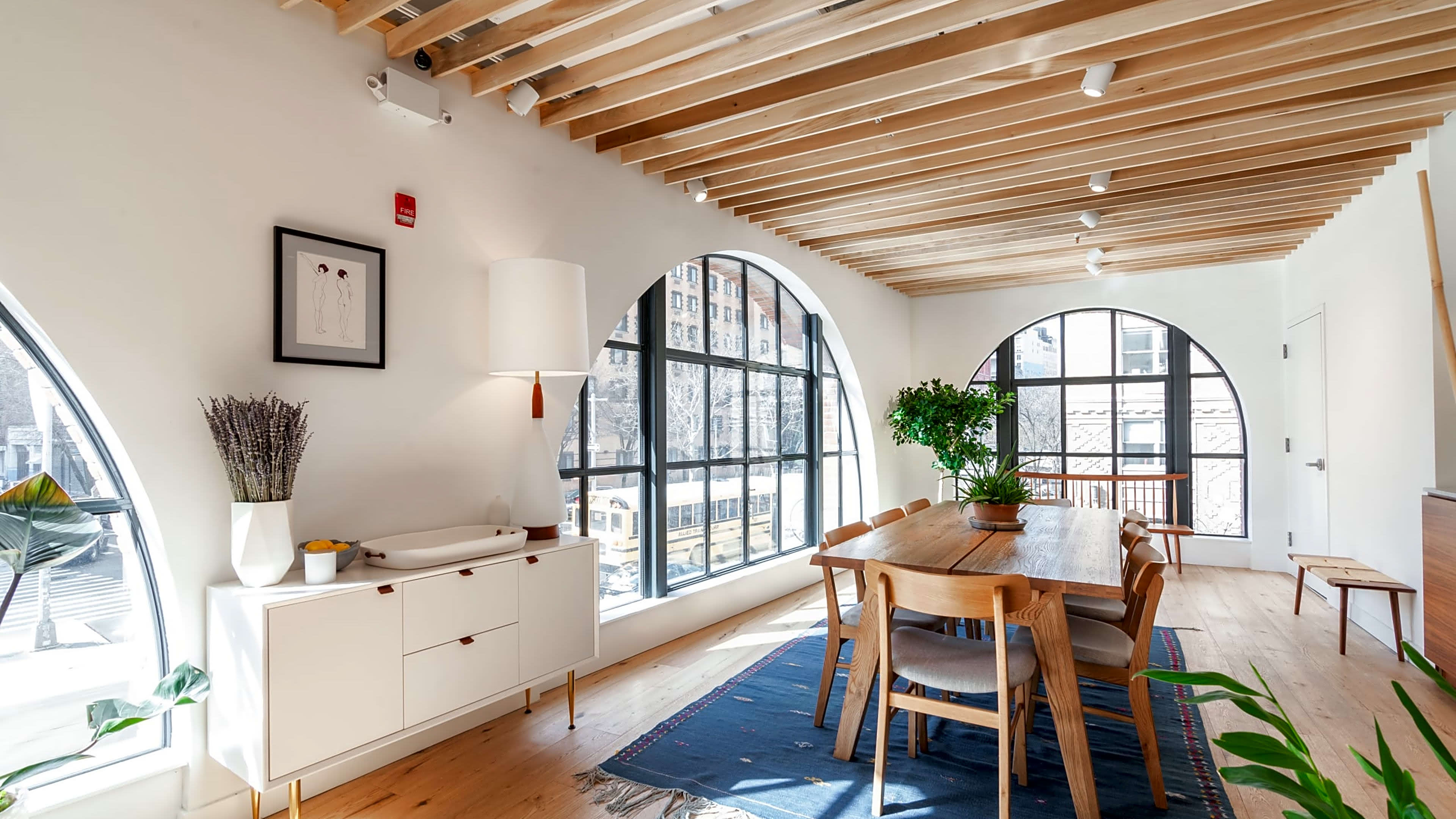 A modern dining area with a wooden table surrounded by chairs, large windows featuring arched designs, and minimalist decor elements.