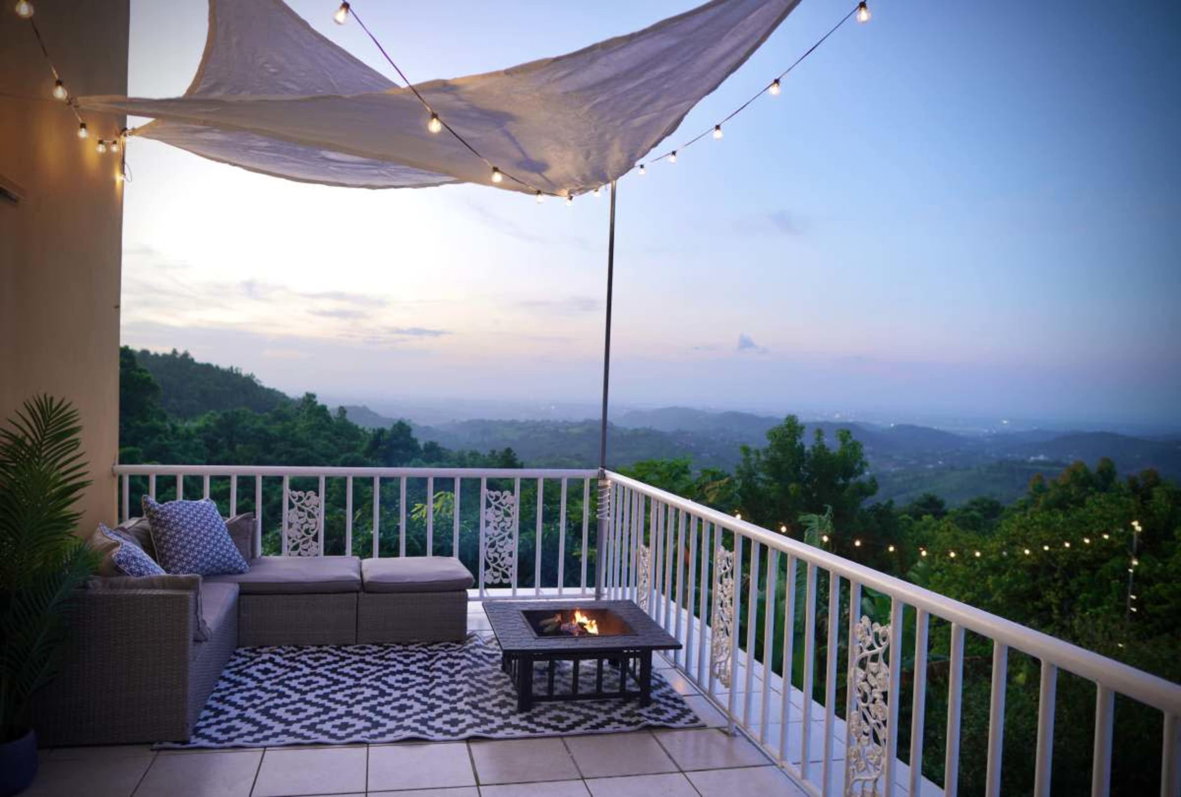 A covered balcony features a seating area with a small fire pit and string lights overlooking a mountainous landscape at dusk.