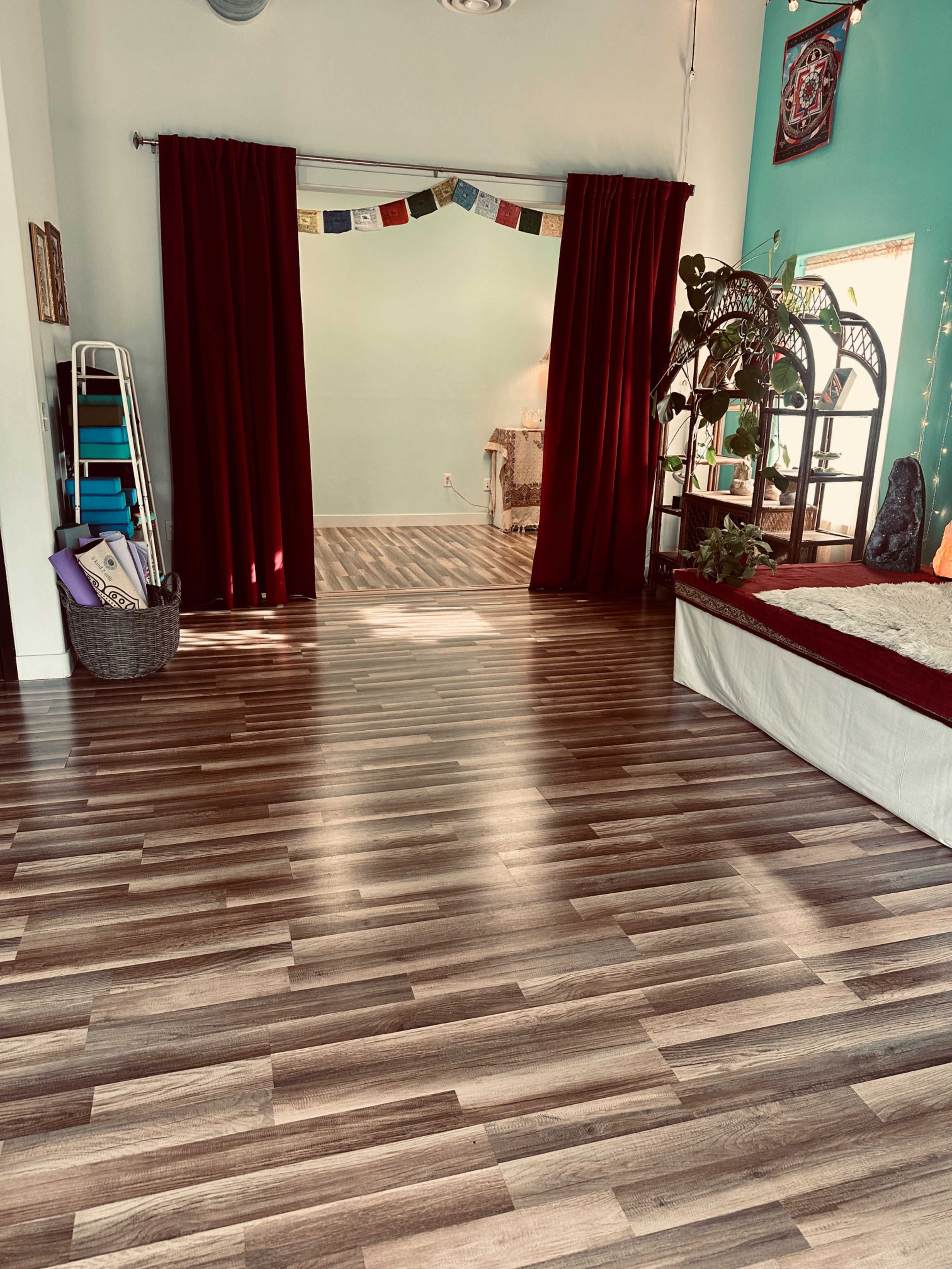 The image shows a room with wooden flooring, red curtains framing an open doorway, a bed with white bedding, and a decorative plant in the corner.