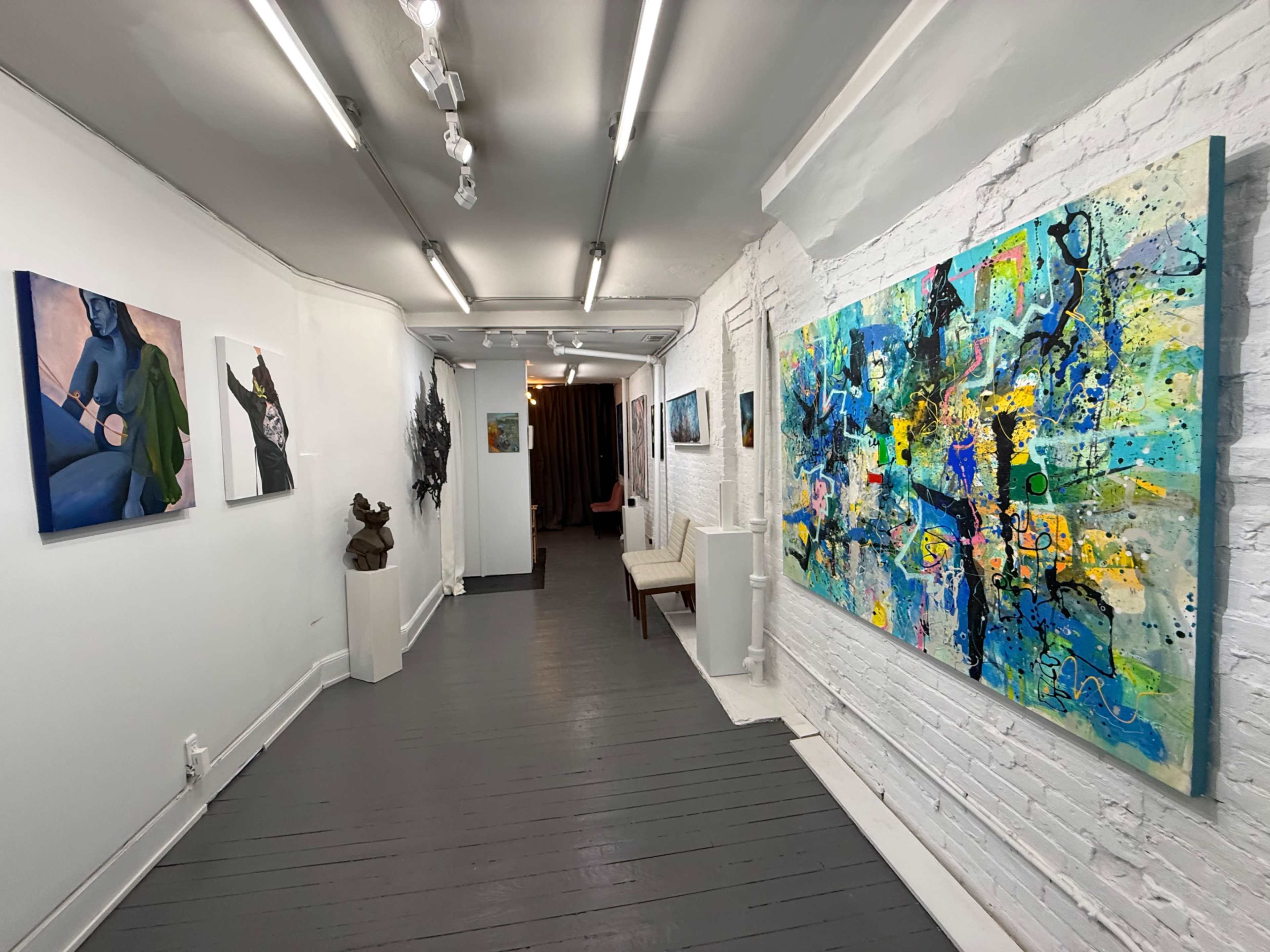 The image shows a gallery hallway featuring various artworks on the walls, including paintings and a sculpture, with a seating area visible in the background.