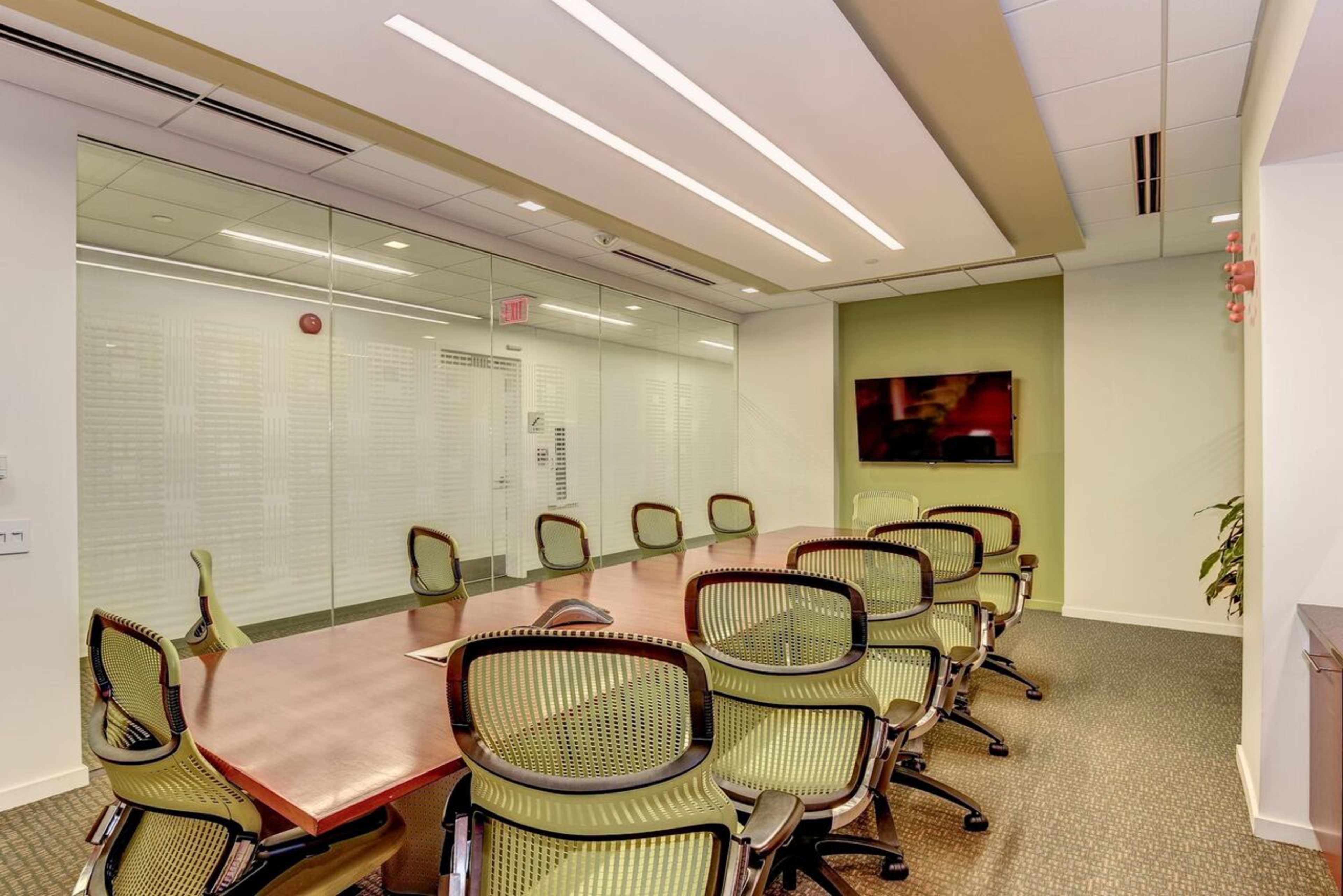 A conference room features a large wooden table surrounded by green ergonomic chairs, with glass walls and a mounted television.