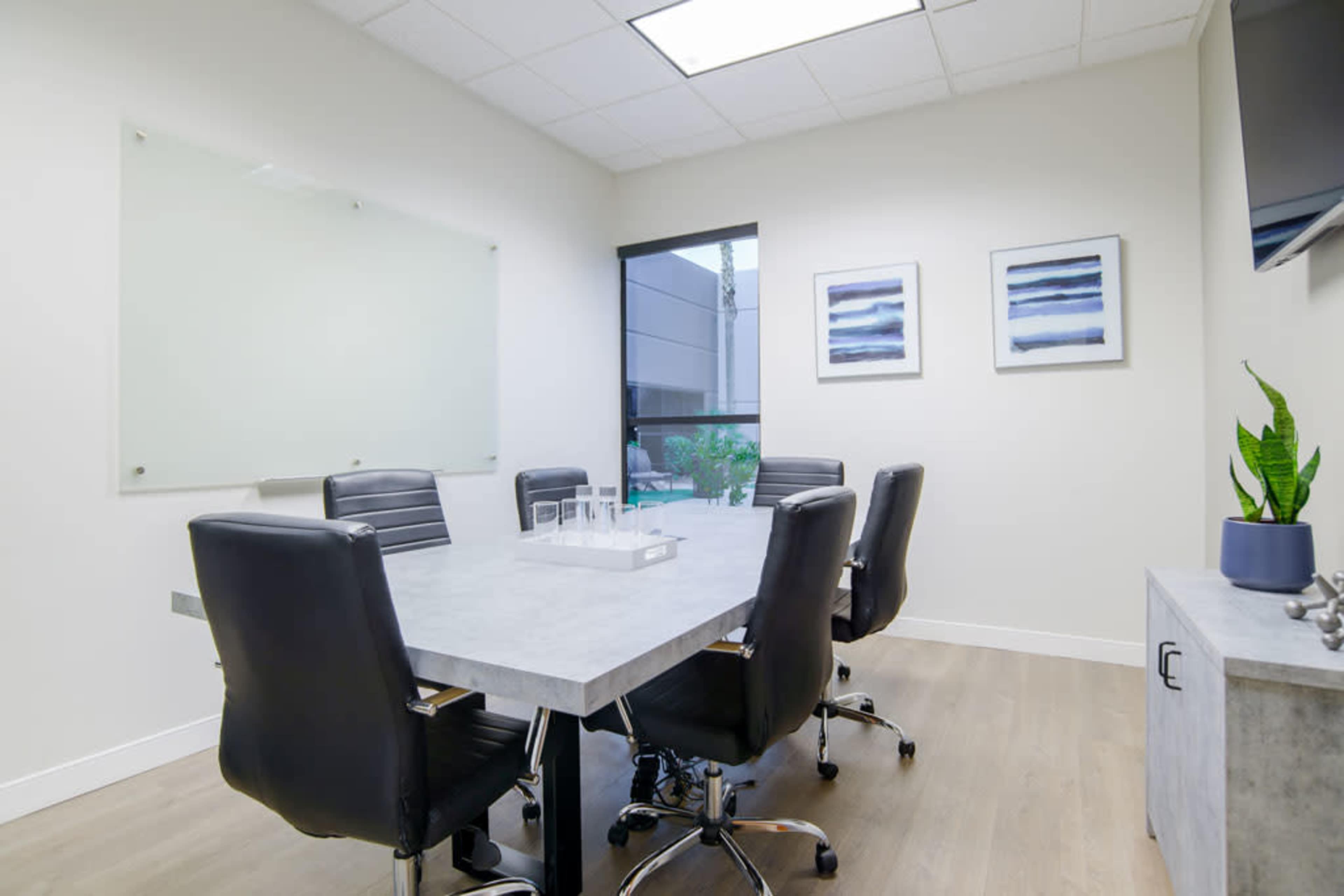 A modern conference room with a large table surrounded by black chairs, a glass whiteboard, and two framed pictures on the wall.
