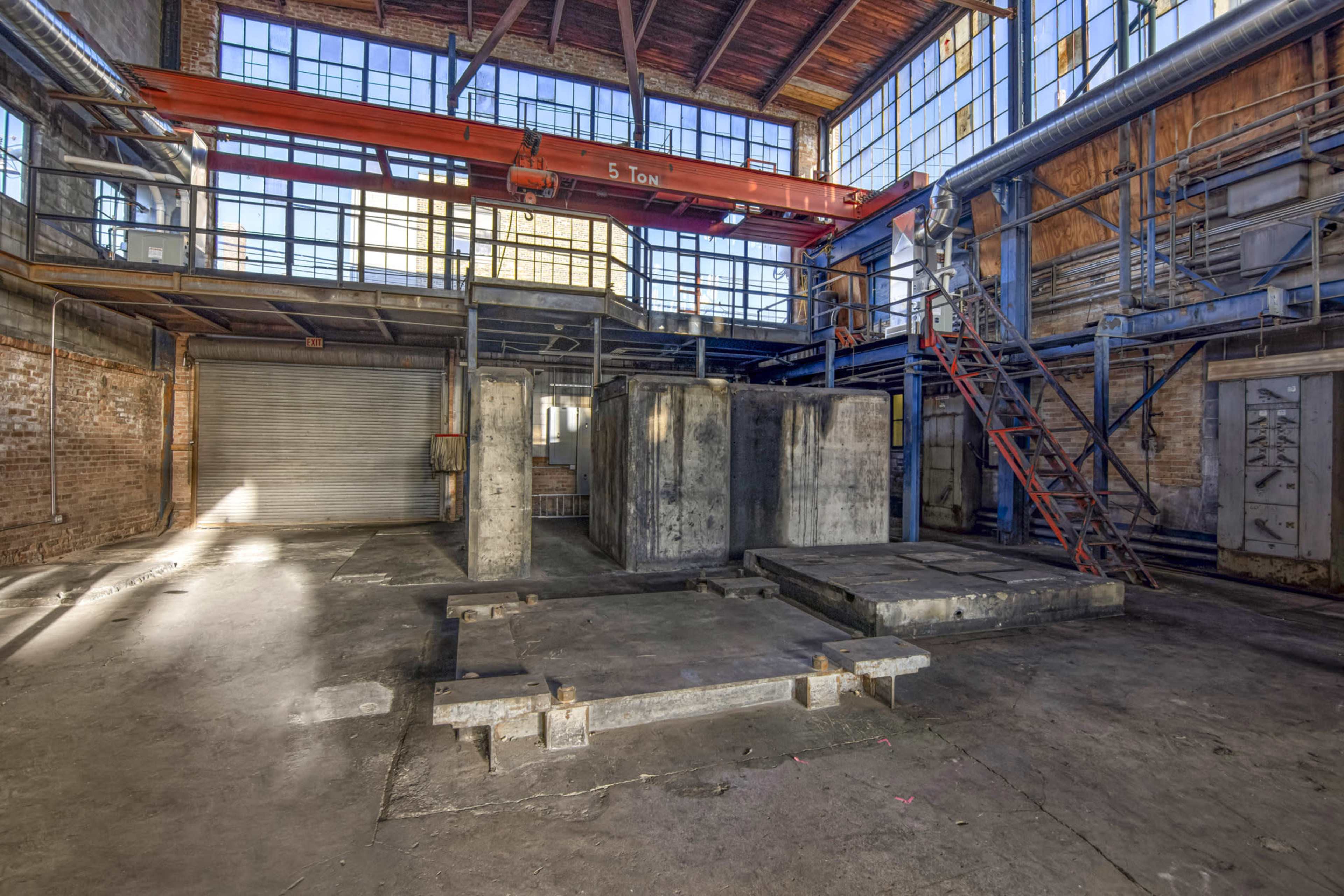 An industrial warehouse interior with concrete structures, a large overhead crane, and a metal staircase leading up to a platform.