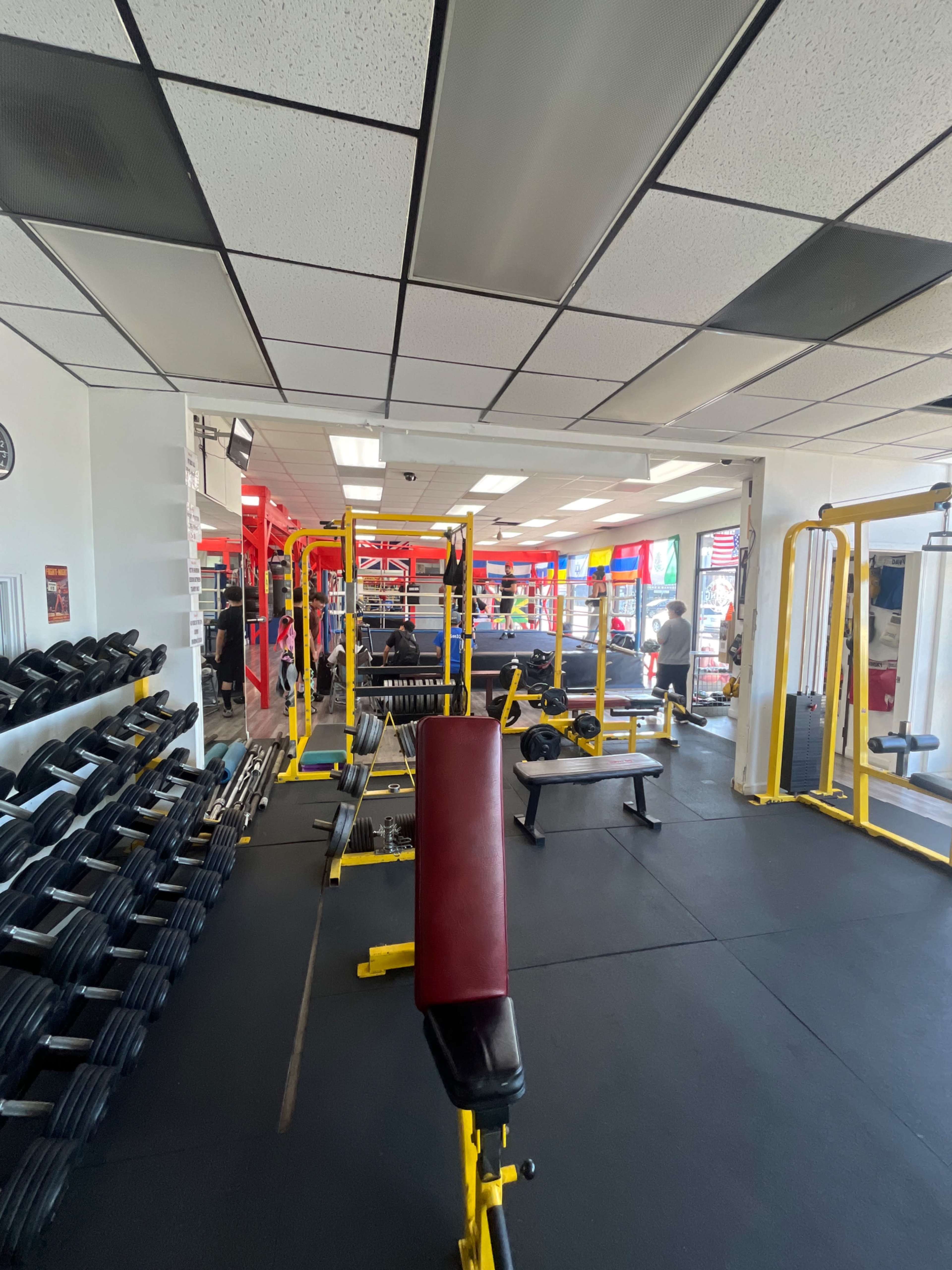 The image shows a gym interior with weightlifting equipment, benches, and multiple exercise stations.