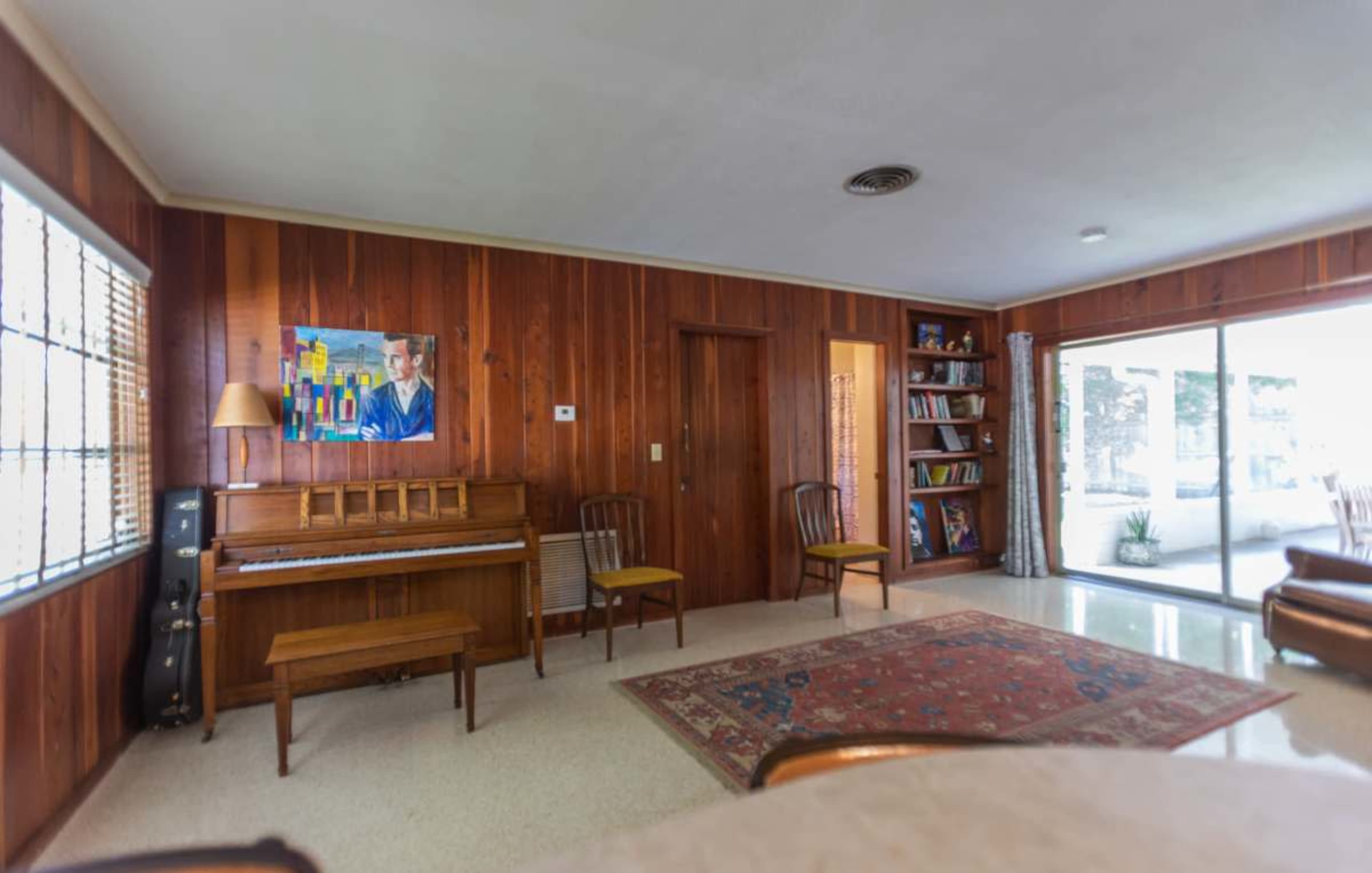 The room features a wooden wall, a piano with a bench, two chairs, a painting, and a large glass door leading to an outdoor area.