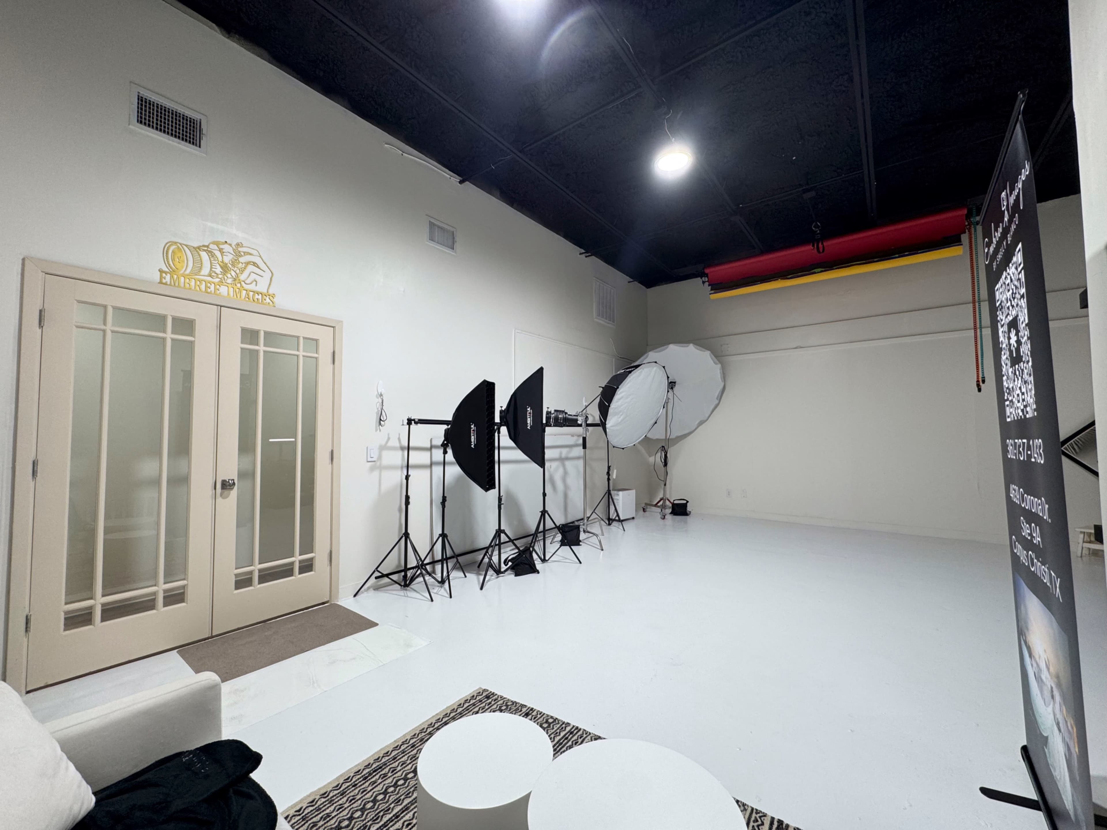 The image shows an empty photography studio with a white floor, two large softbox lights on stands, and a variety of backdrops suspended from the ceiling.