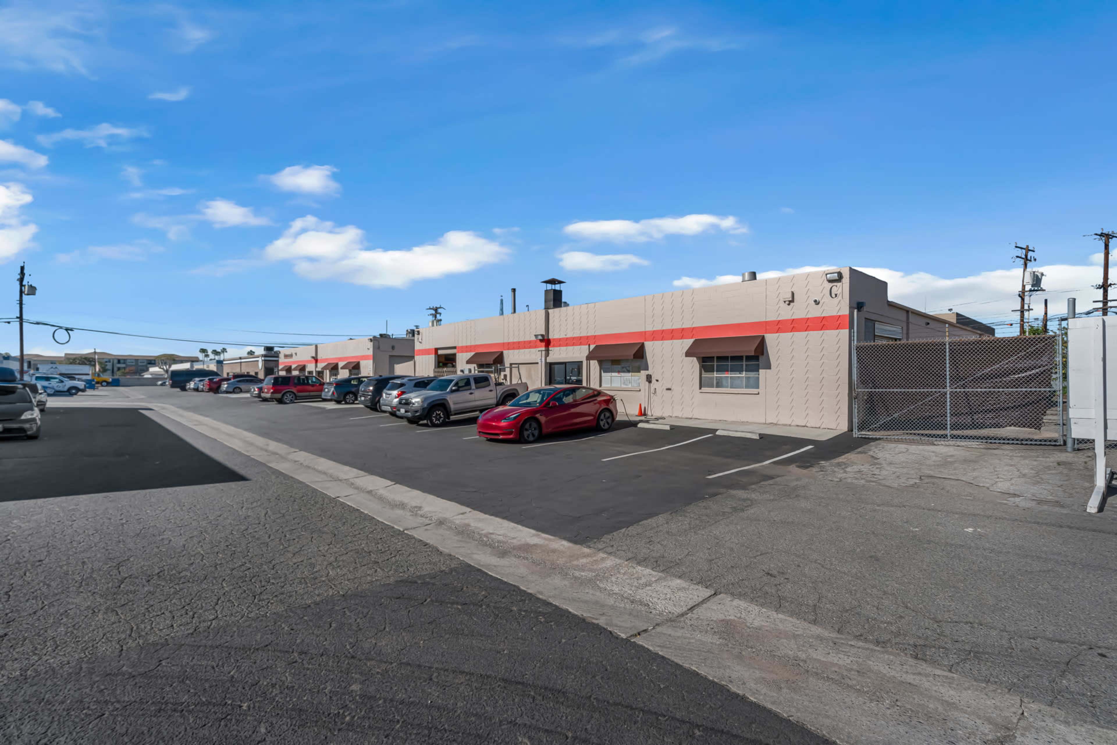 The image shows a row of industrial buildings with parked cars in front, set against a clear blue sky.