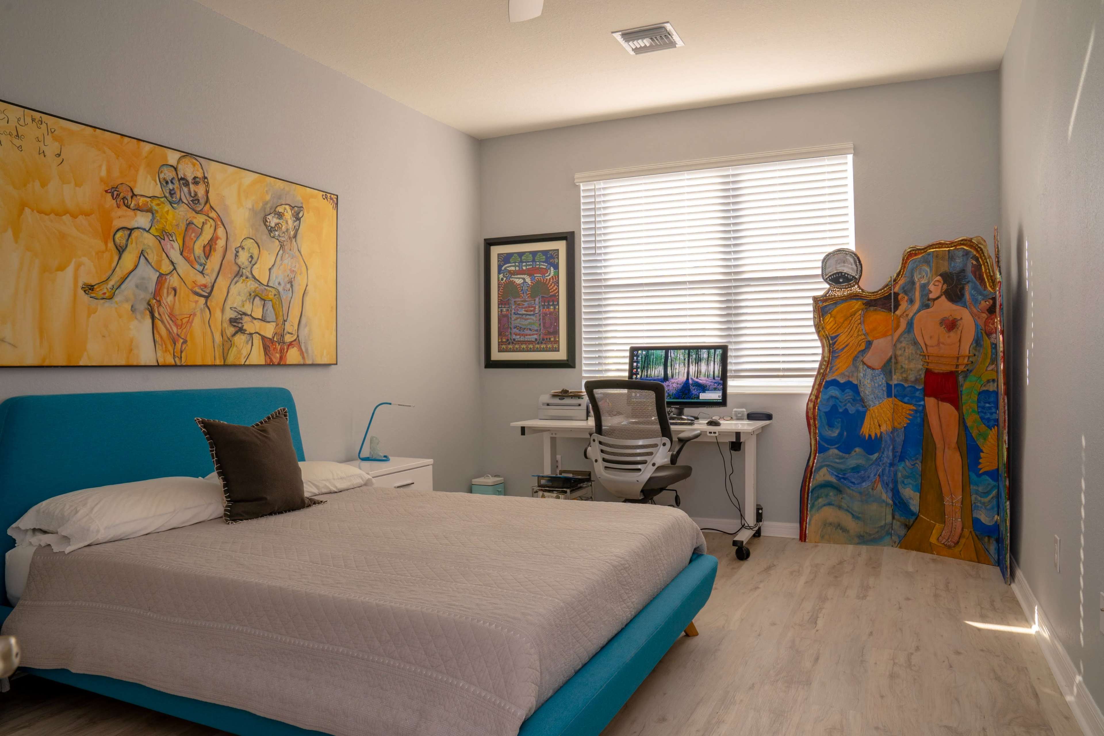 A minimalist bedroom features a blue bed with a light-colored blanket, a small nightstand, a desk with a computer, and colorful artwork on the walls.