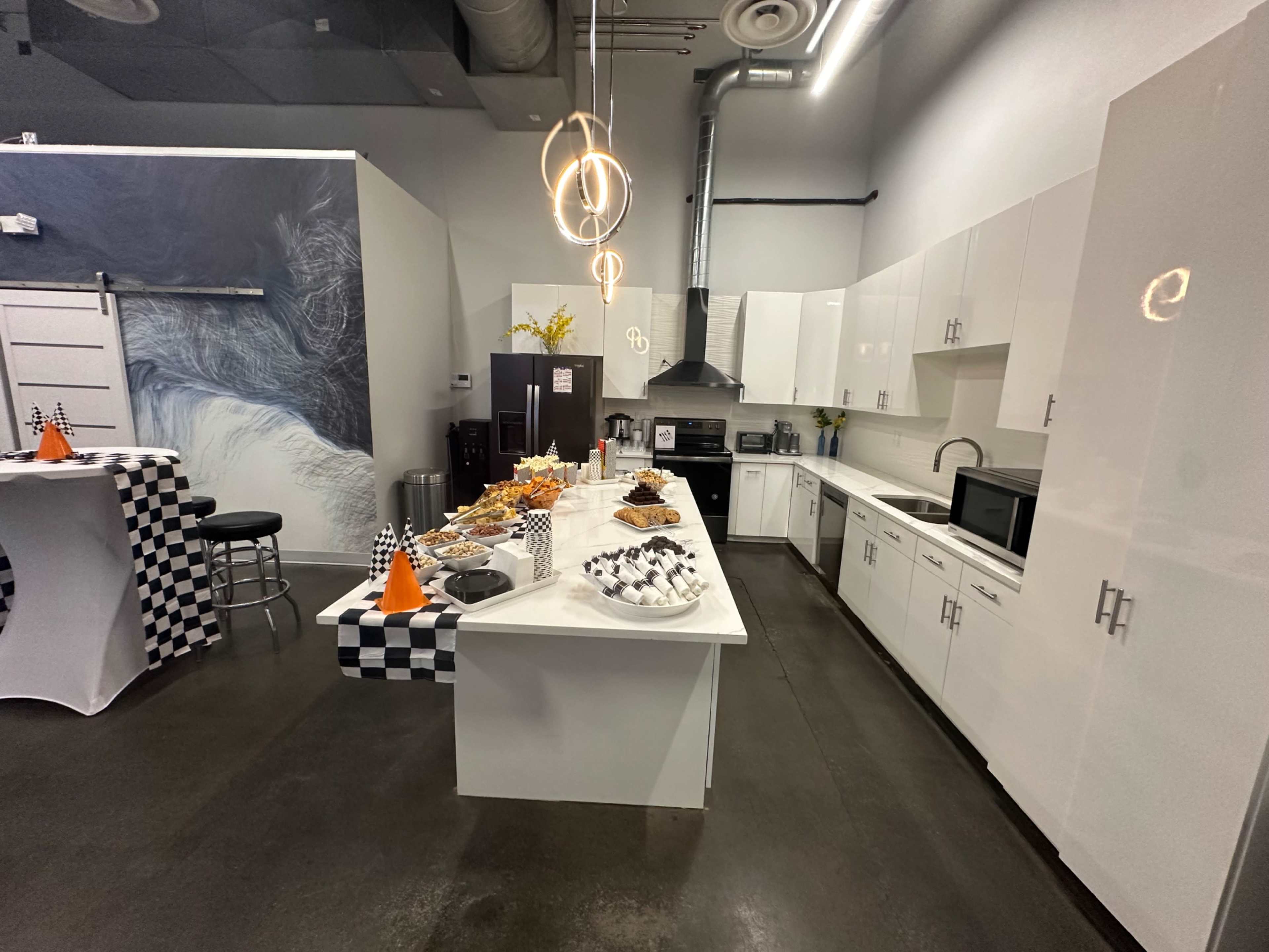 The image shows a modern kitchen area with white cabinets, a central island displaying food, and a checkered tablecloth on a nearby table.