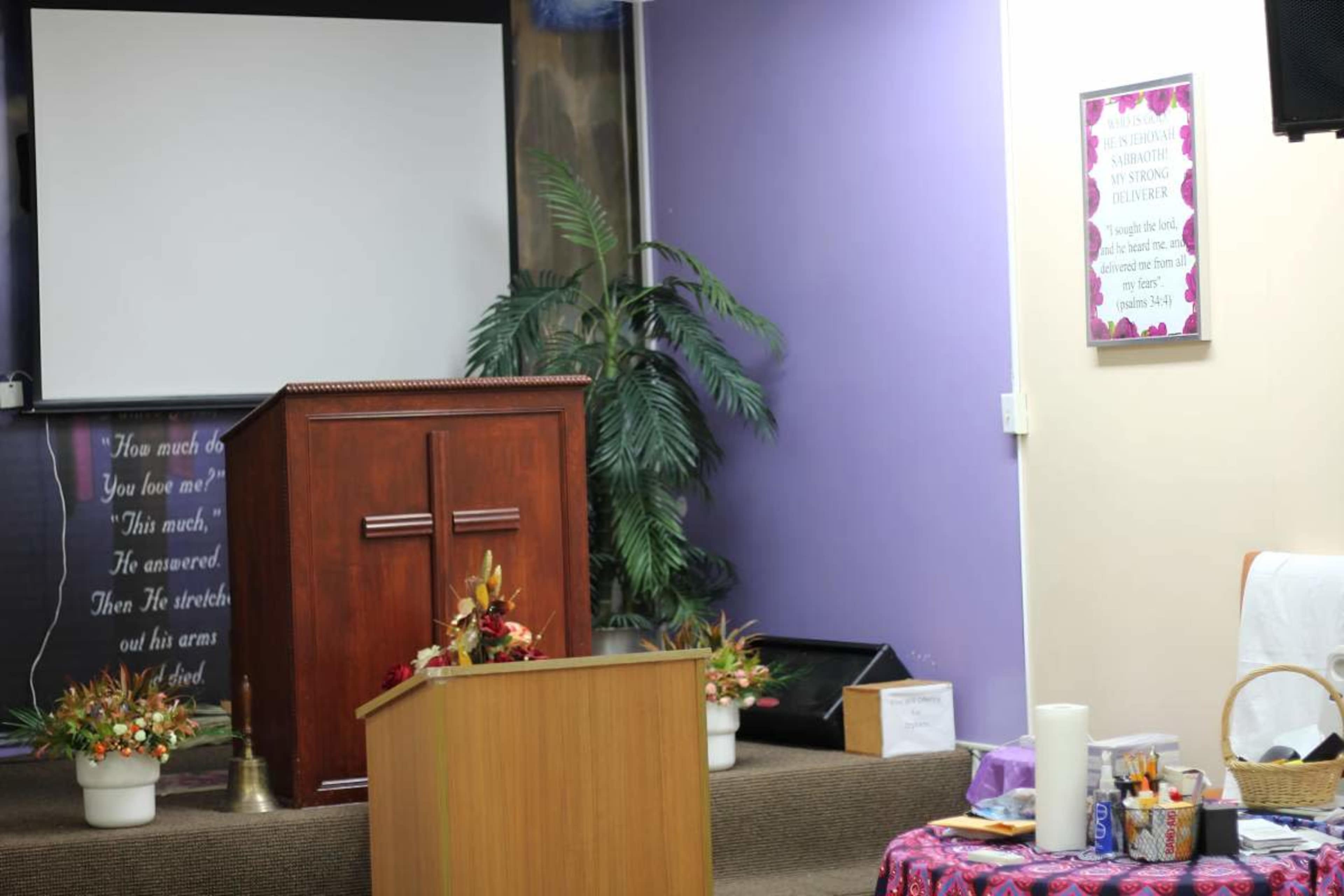 The image shows a modest interior of a community space or church, featuring a wooden podium, a projector screen, decorative plants, and a table with various items on it.