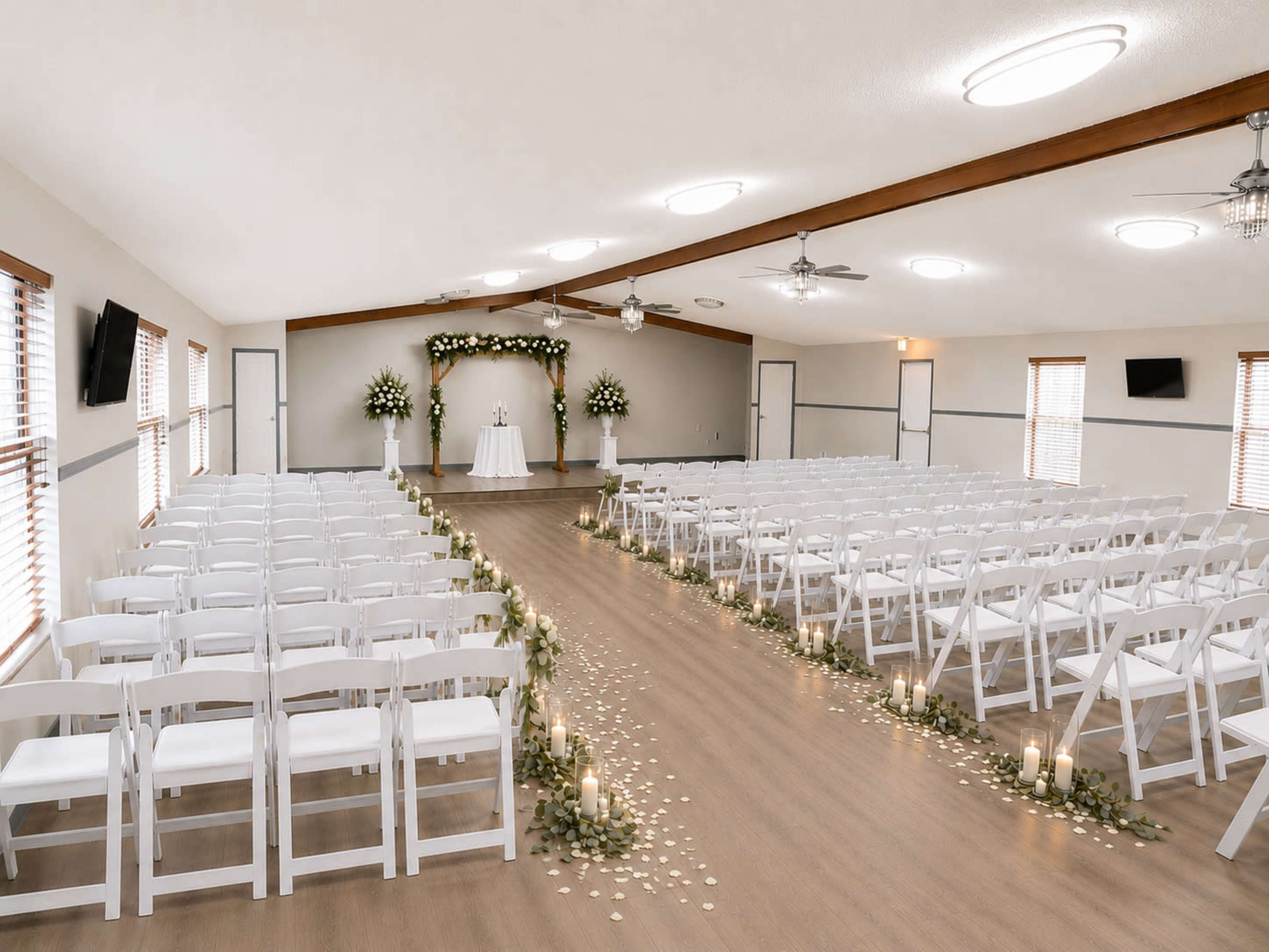The image shows an indoor wedding ceremony setup with rows of white chairs facing an altar adorned with flowers and candles along a decorated aisle.