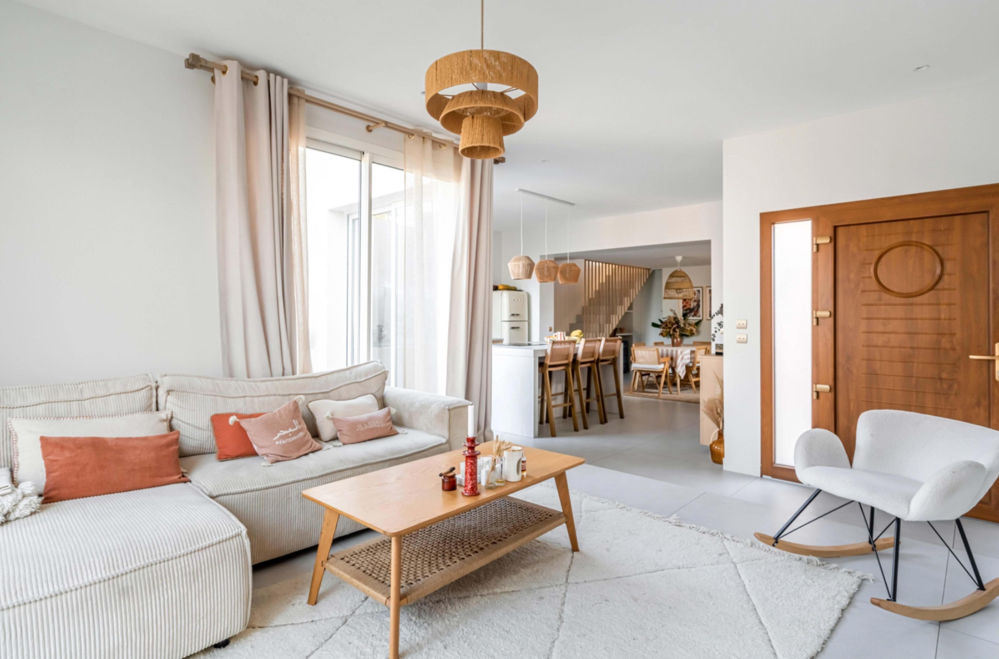 A modern living room features a light-colored sofa with decorative pillows, a wooden coffee table, and a rocking chair, all surrounded by large windows and minimalistic decor.