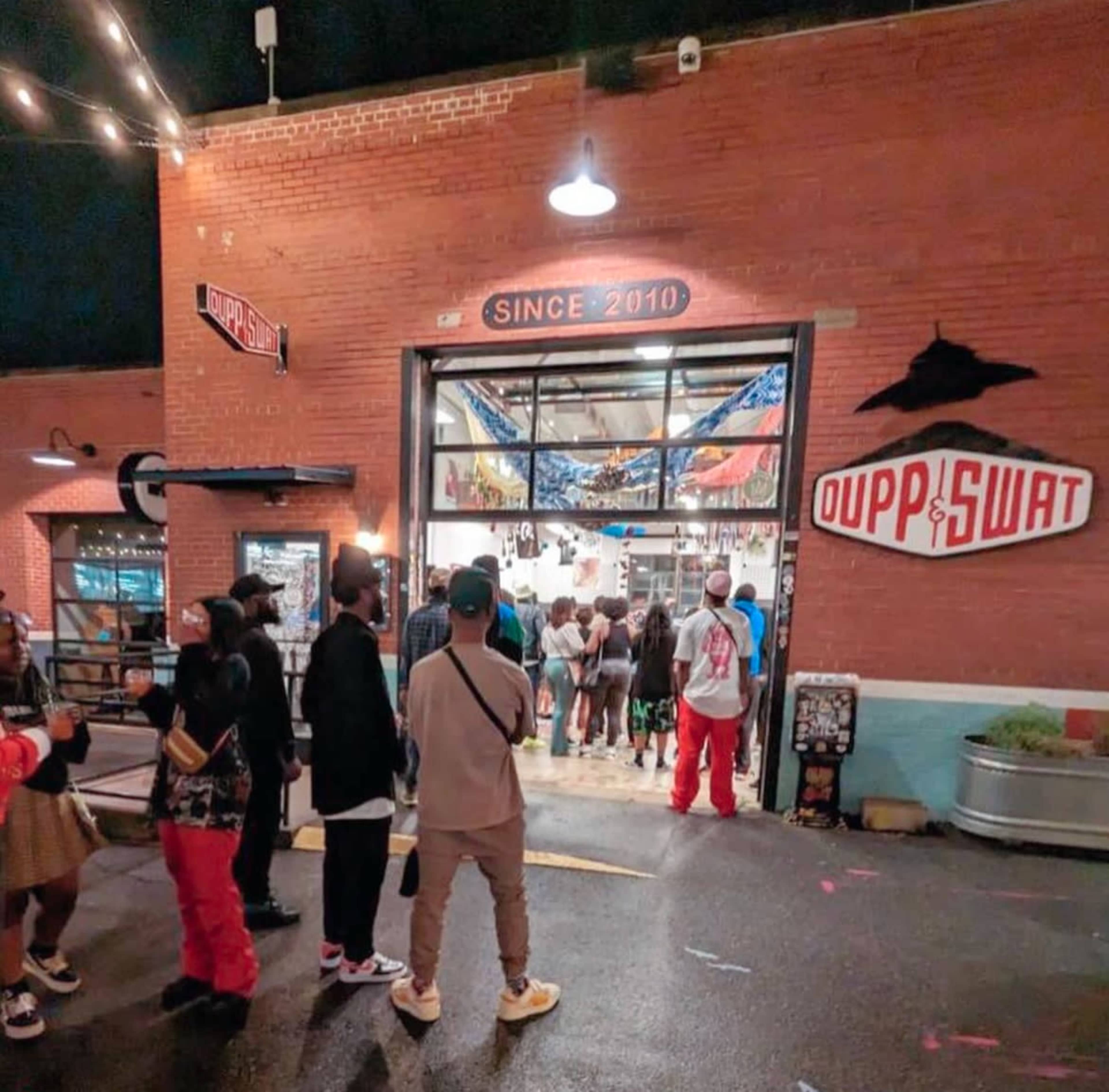 A group of people stands in line outside a store named "UPPERSWAT" beneath a sign indicating it has been established since 2010.