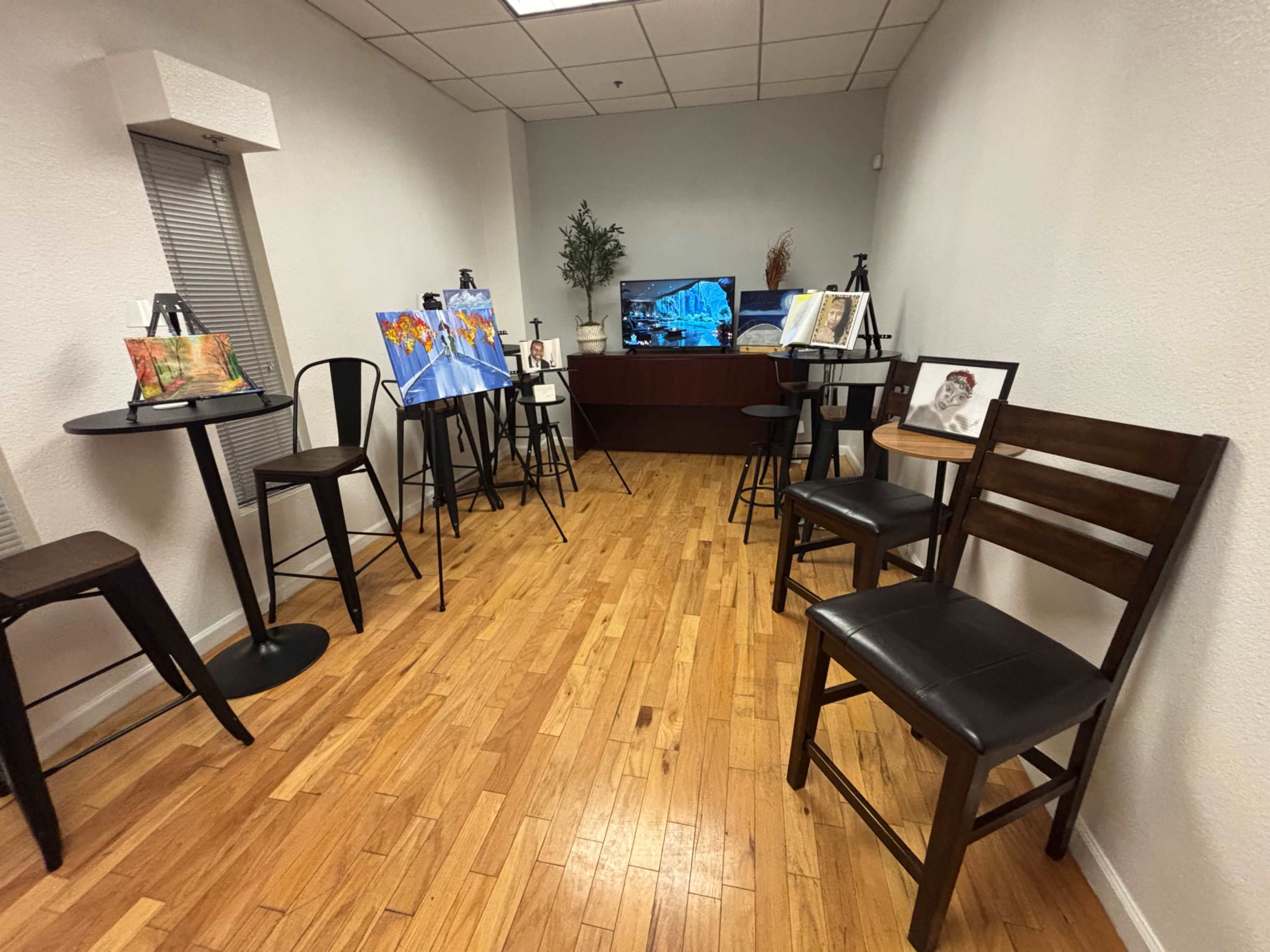 The image shows a small, well-lit room featuring several art pieces displayed on easels, surrounded by a few bar-style tables and chairs.