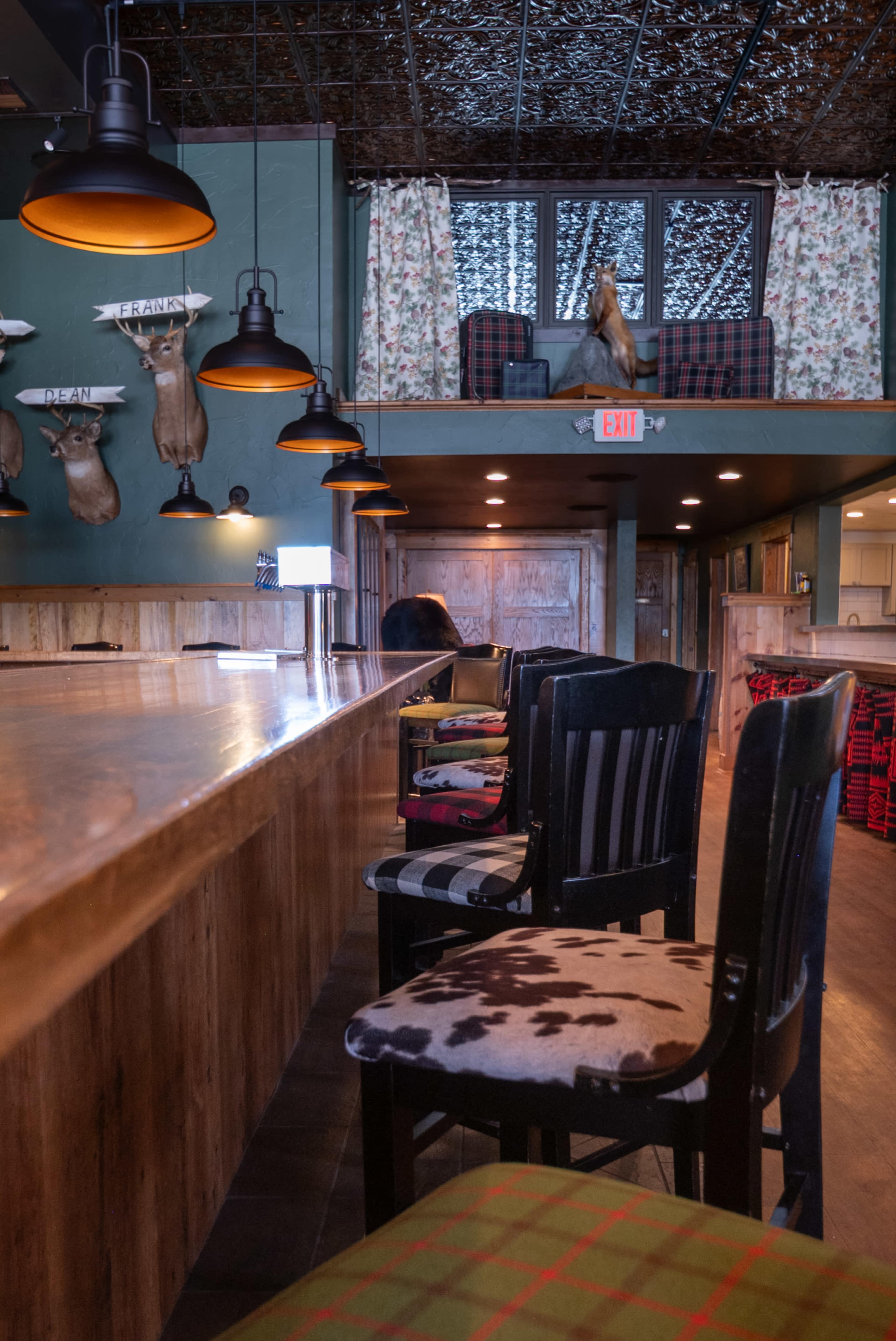 The image shows a bar with a long wooden countertop, several chairs featuring various patterns, and taxidermy mounted on the walls in a rustic setting.