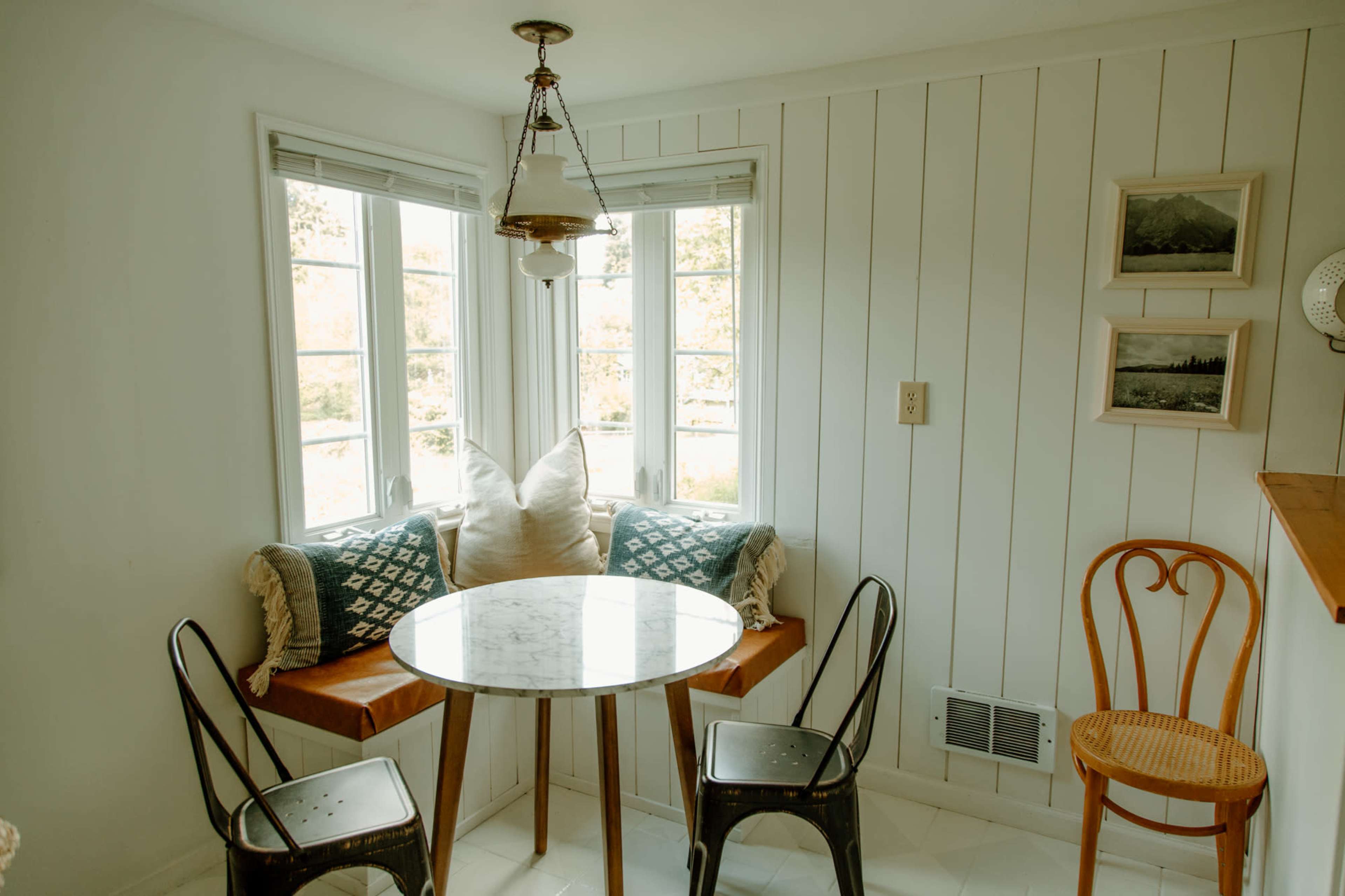 A small dining area features a round marble table surrounded by two chairs, with a built-in window seat adorned with cushions.