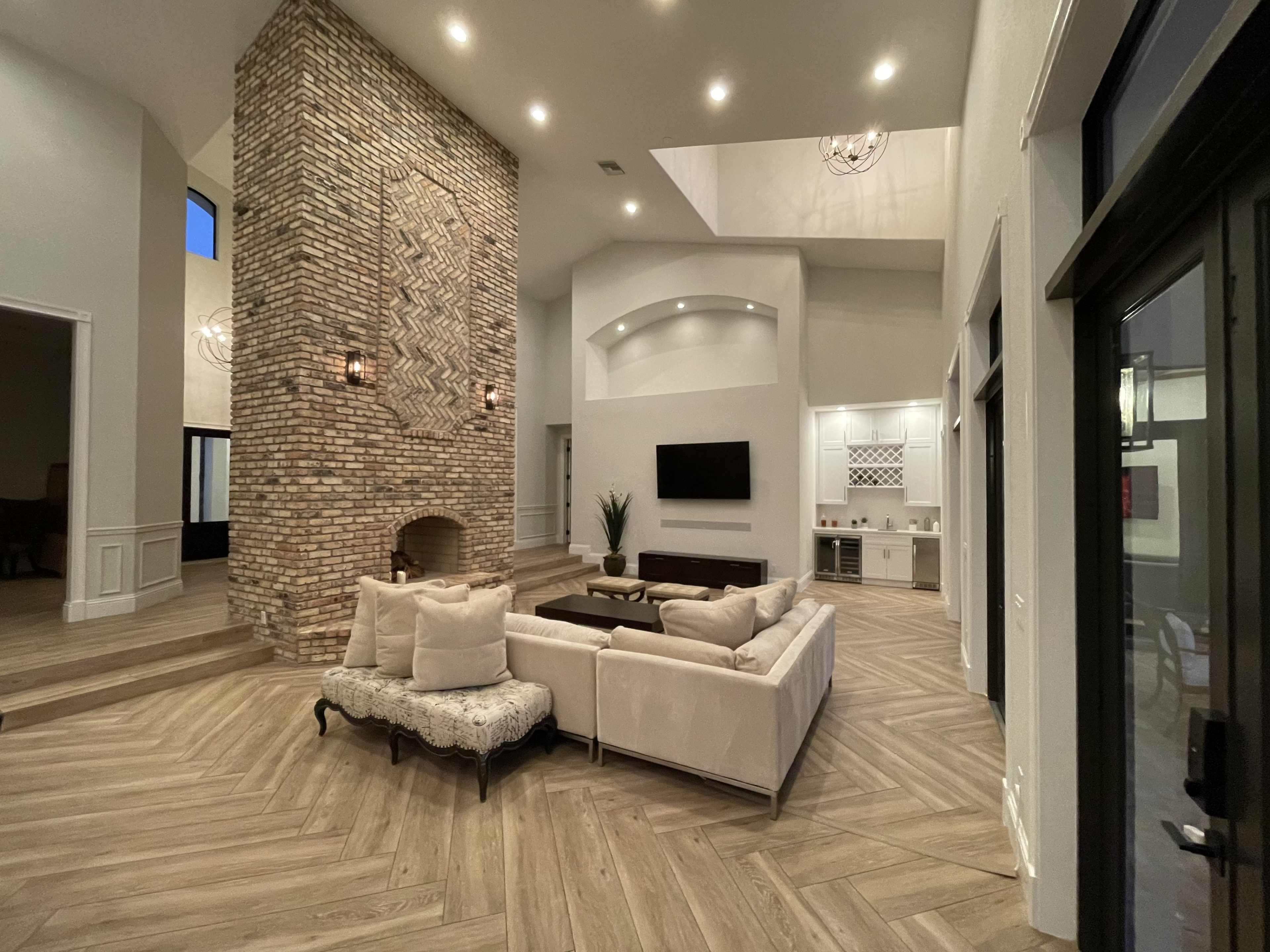 A spacious living area featuring a large brick fireplace, a sectional sofa, and wooden flooring arranged in a herringbone pattern.