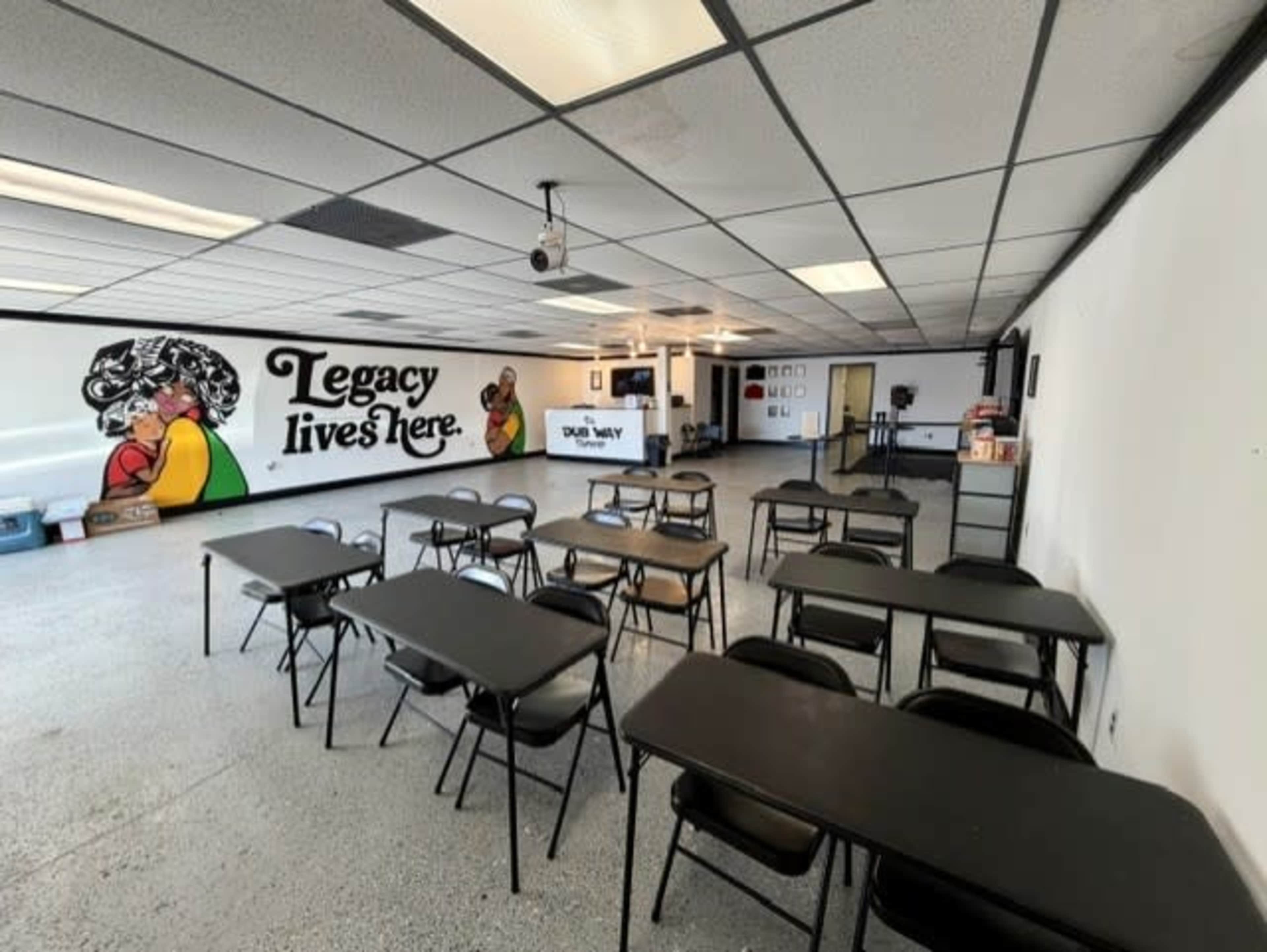 The image shows a classroom setup with several black folding tables and chairs arranged in rows, and a mural on the wall that reads "Legacy lives here."