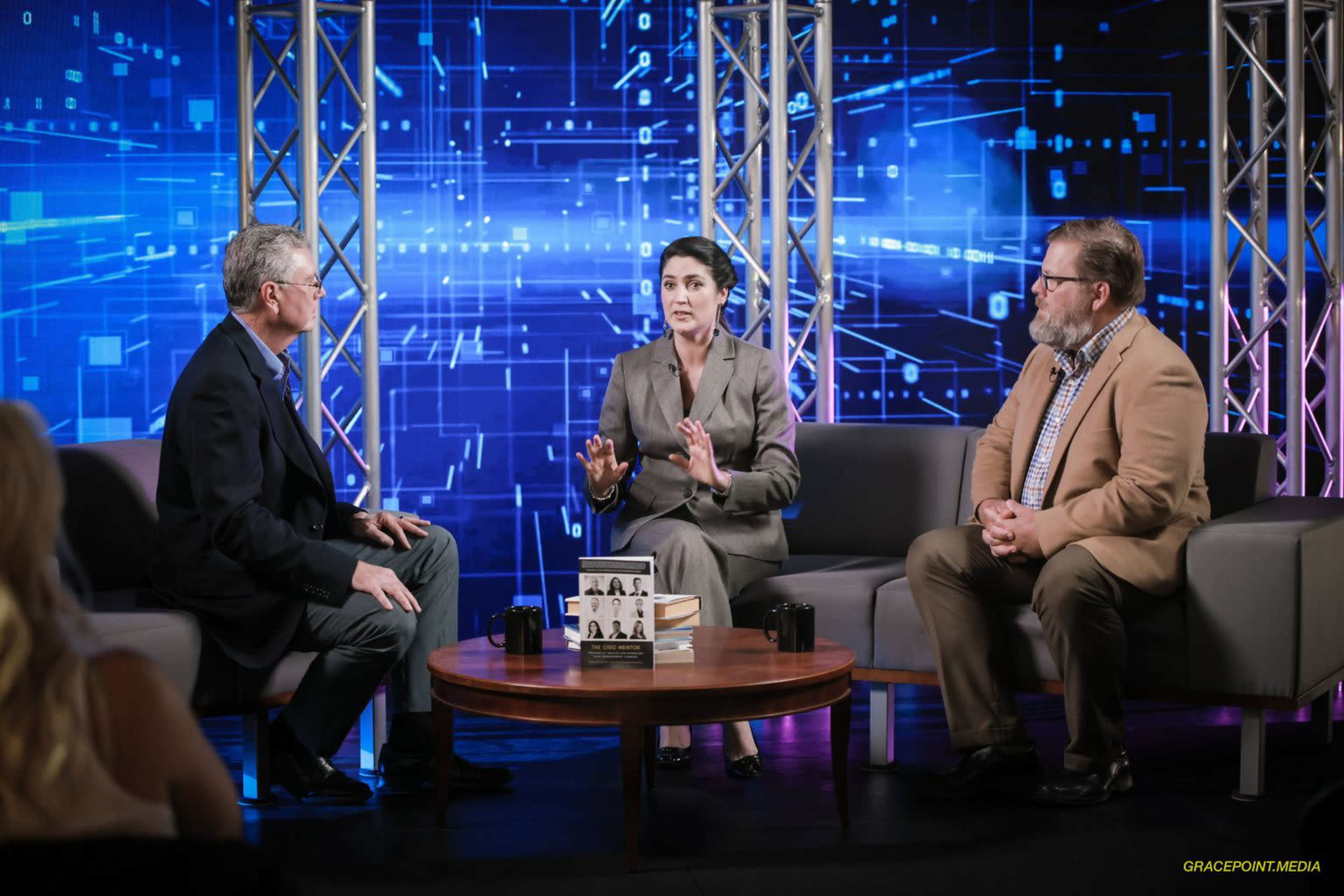 Three people engaged in a discussion on a television set with a digital backdrop featuring blue and purple graphics.
