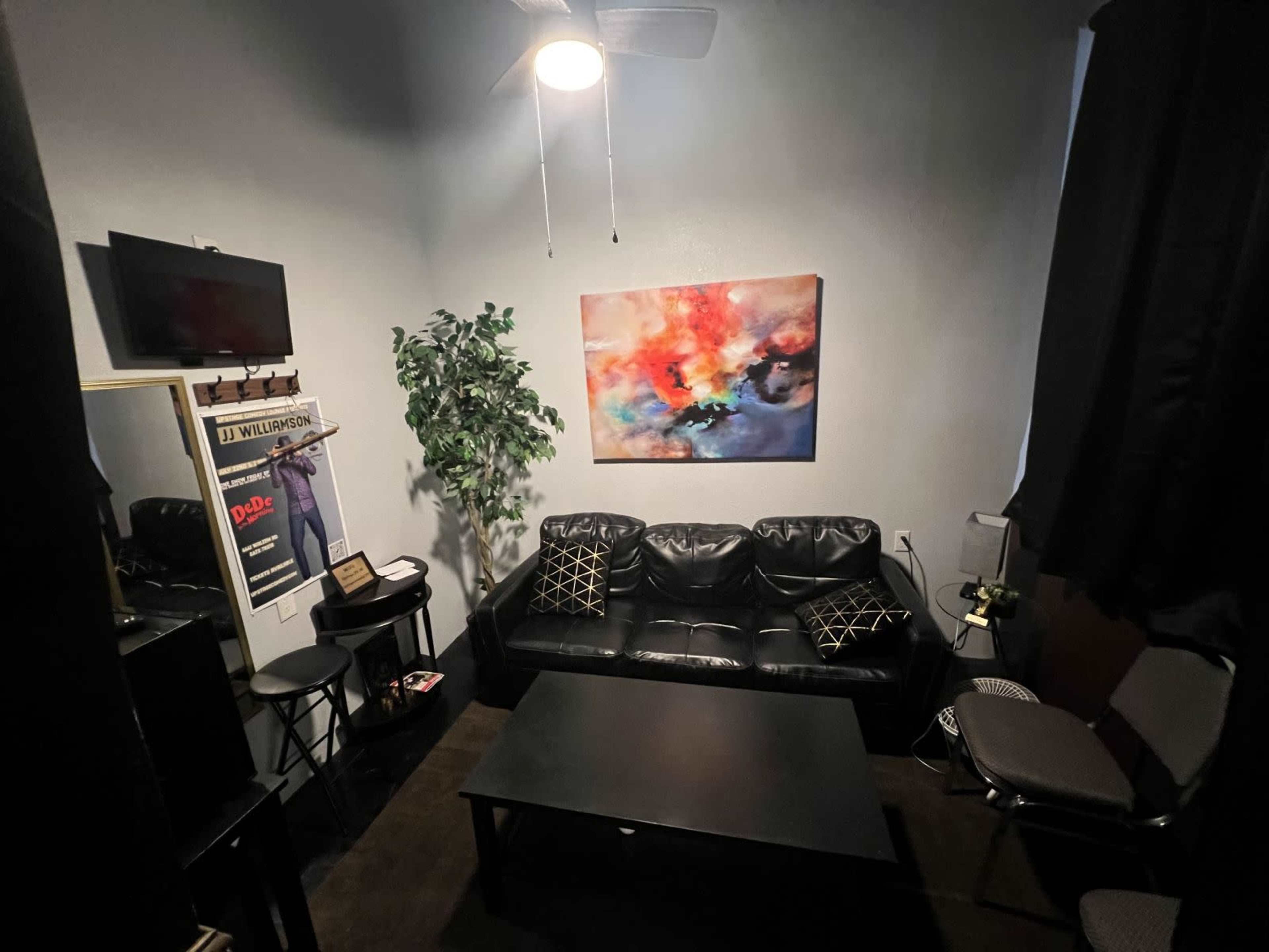 A small room featuring a black leather couch, a coffee table, a decorative plant, a wall-mounted TV, and an abstract painting.