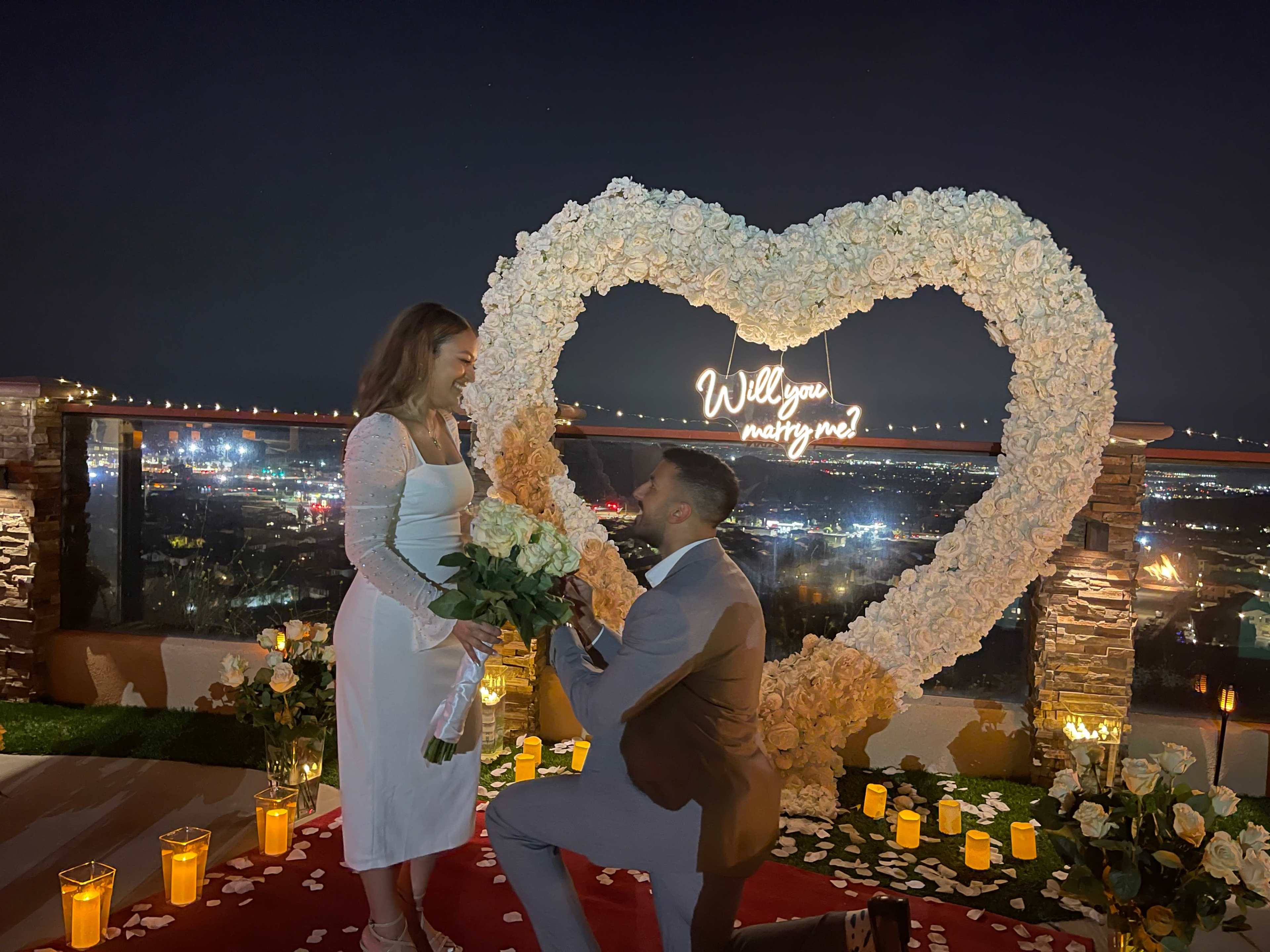 A man is proposing to a woman in front of a large heart-shaped flower arrangement illuminated with the words "Will you marry me?" at a nighttime rooftop setting.
