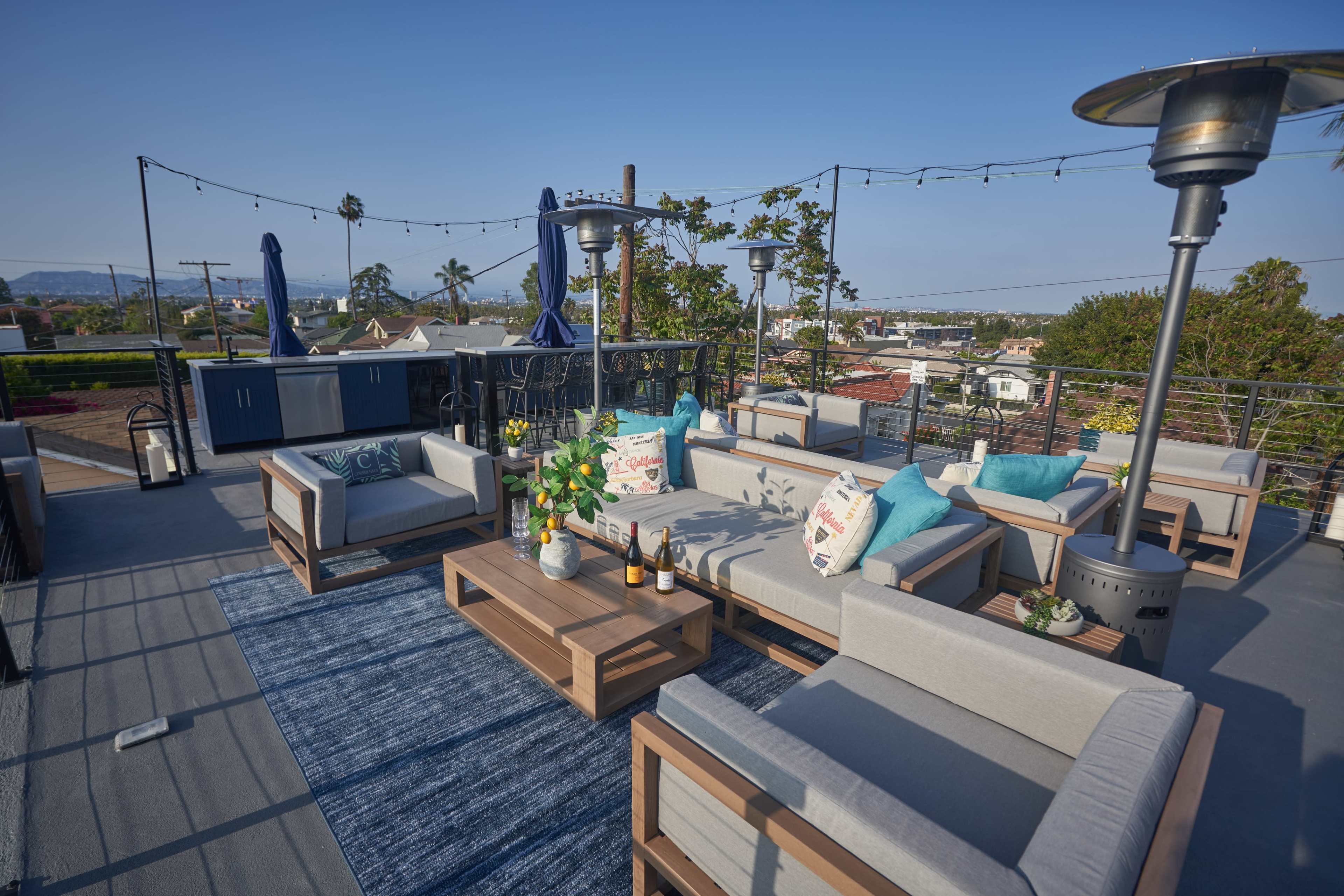 The rooftop deck features modern seating, a table with drinks, and a view of surrounding houses and palm trees.