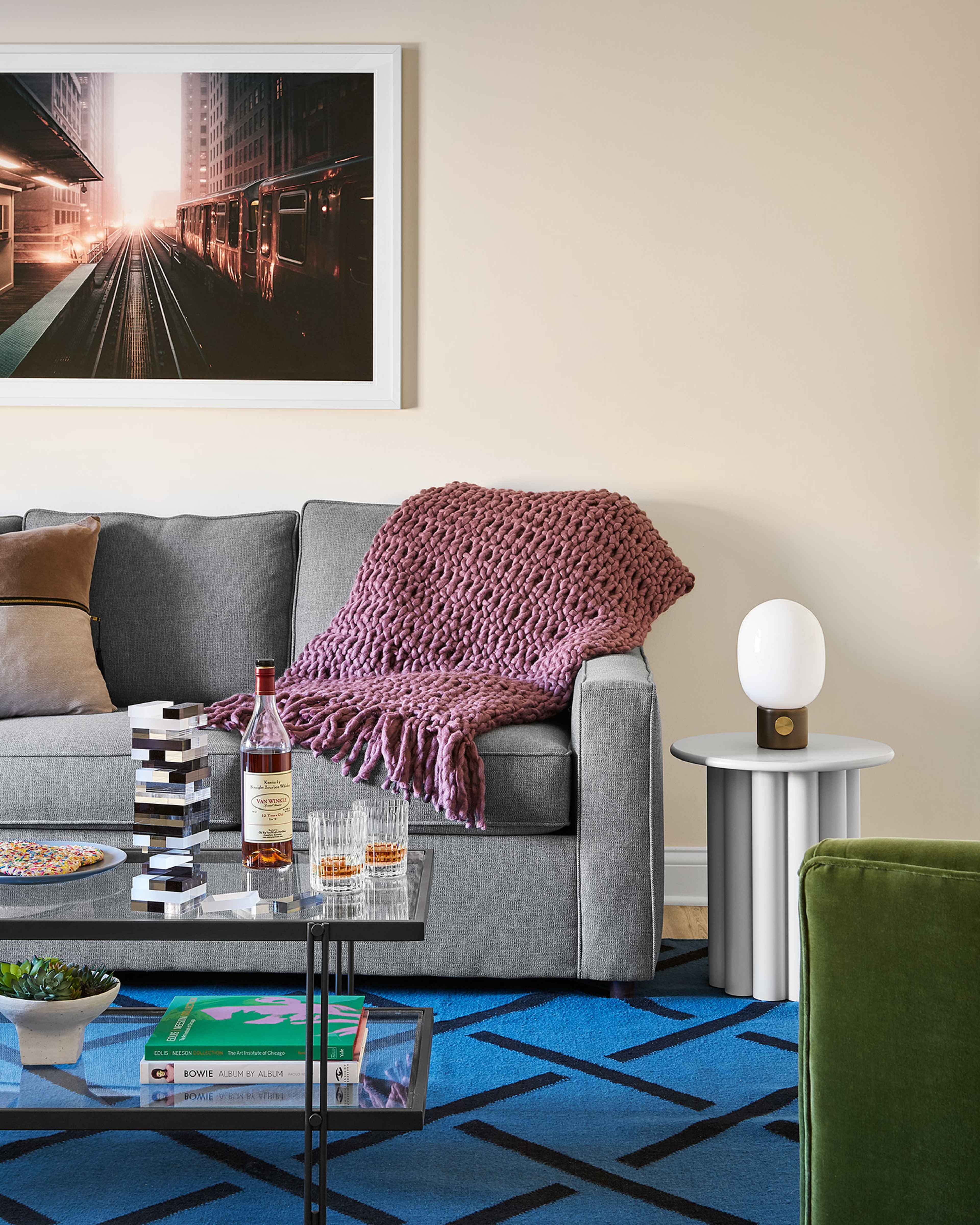 A modern living room features a grey couch adorned with a knitted blanket, alongside a glass coffee table and a decorative side table.