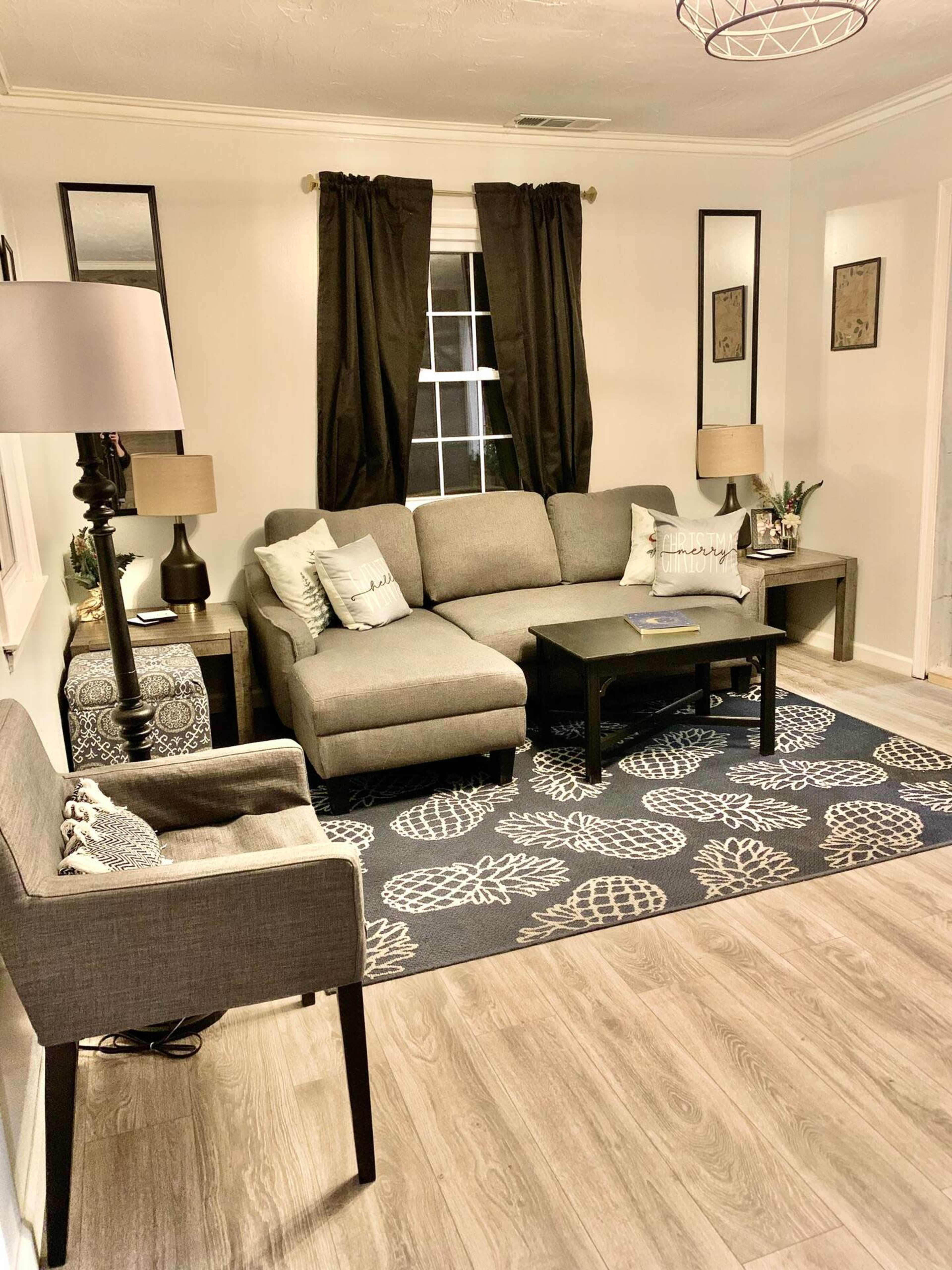 A cozy living room featuring a gray sectional sofa, a black coffee table, and a patterned area rug, with black curtains framing a window.