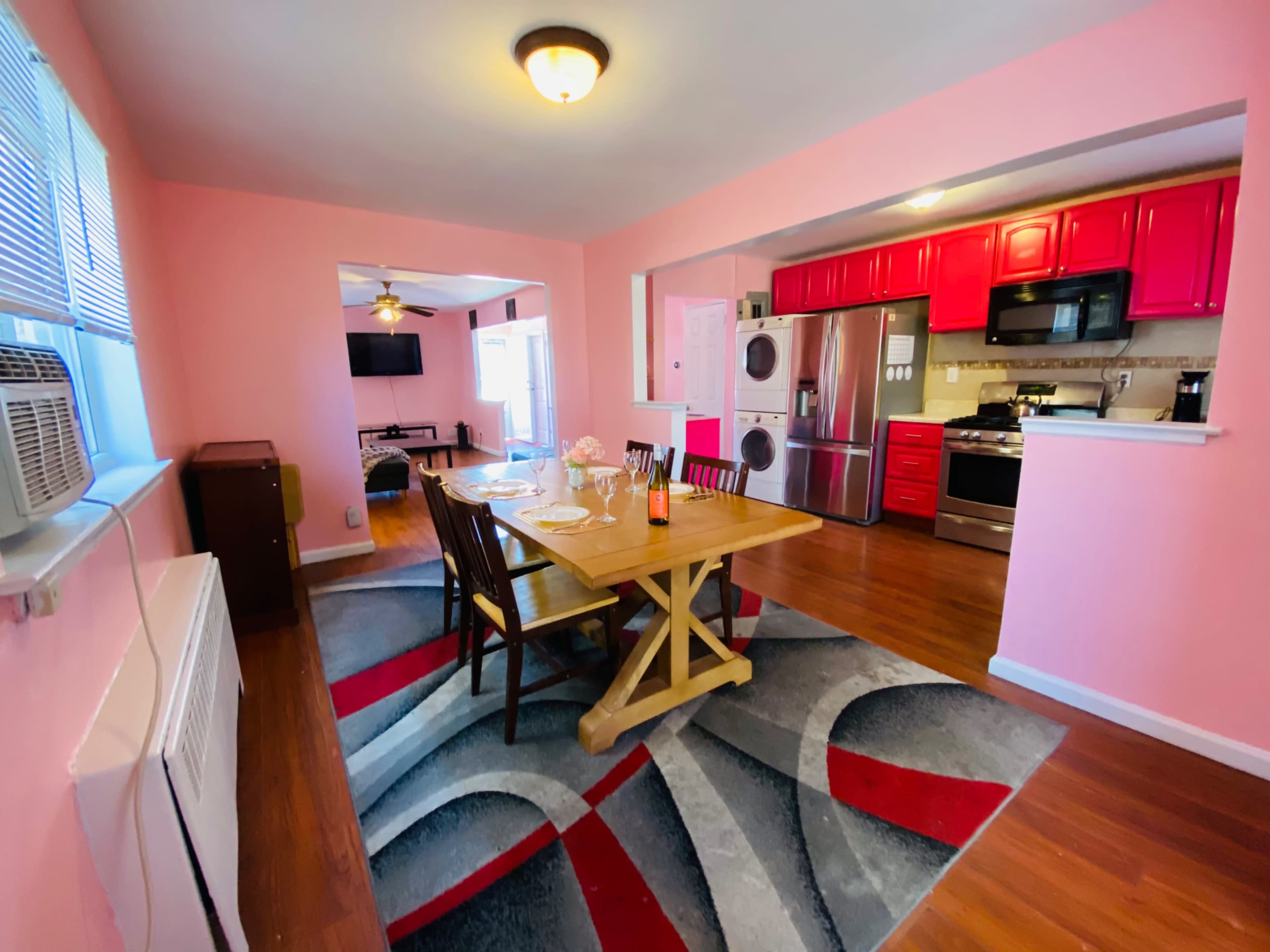 A dining area with a wooden table set for a meal, surrounded by pink walls and leading into a kitchen with red cabinets and stainless steel appliances.