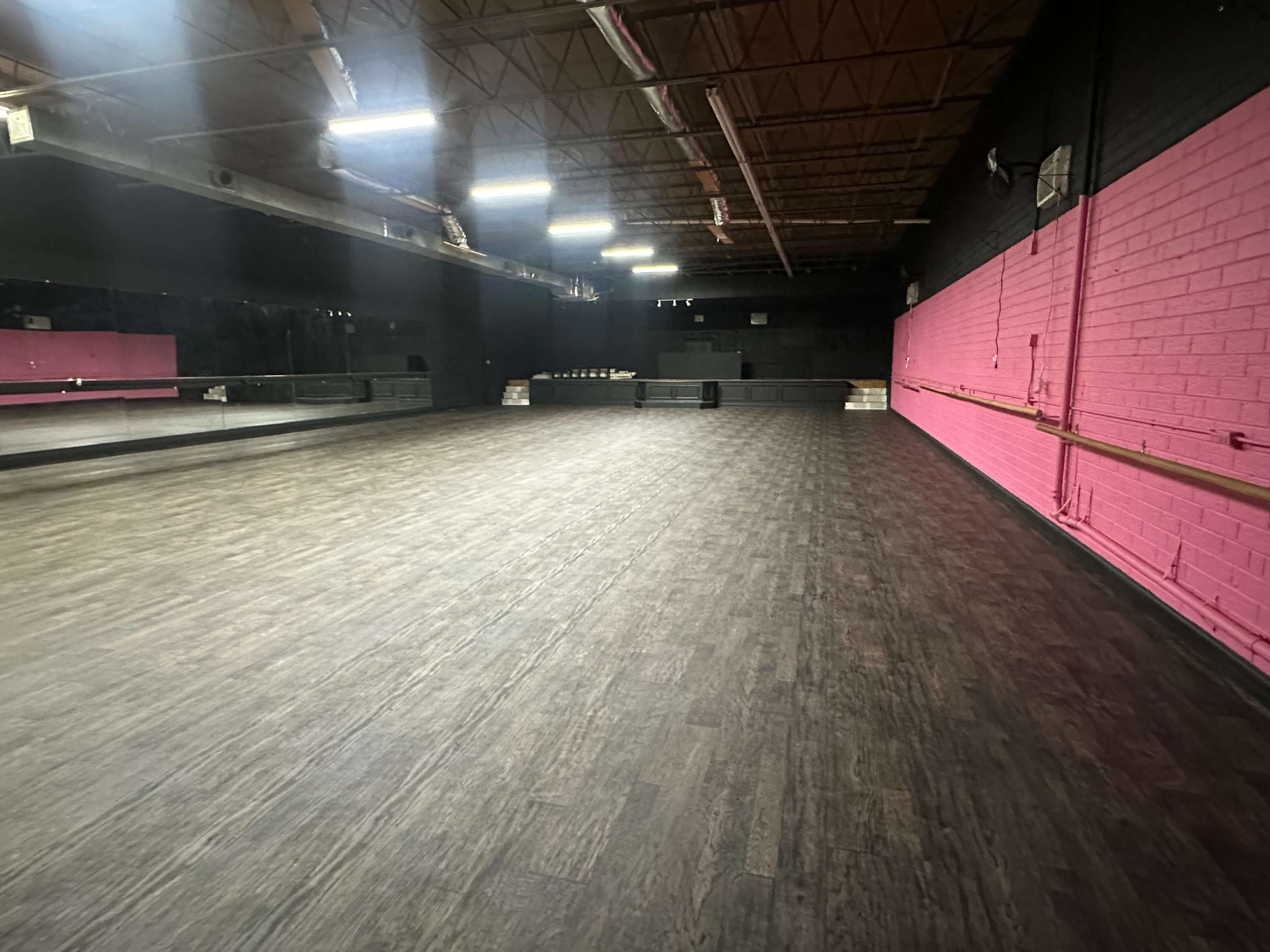 The image shows an empty dance studio with a wooden floor and black walls, featuring a pink accent wall and mirrored panels.