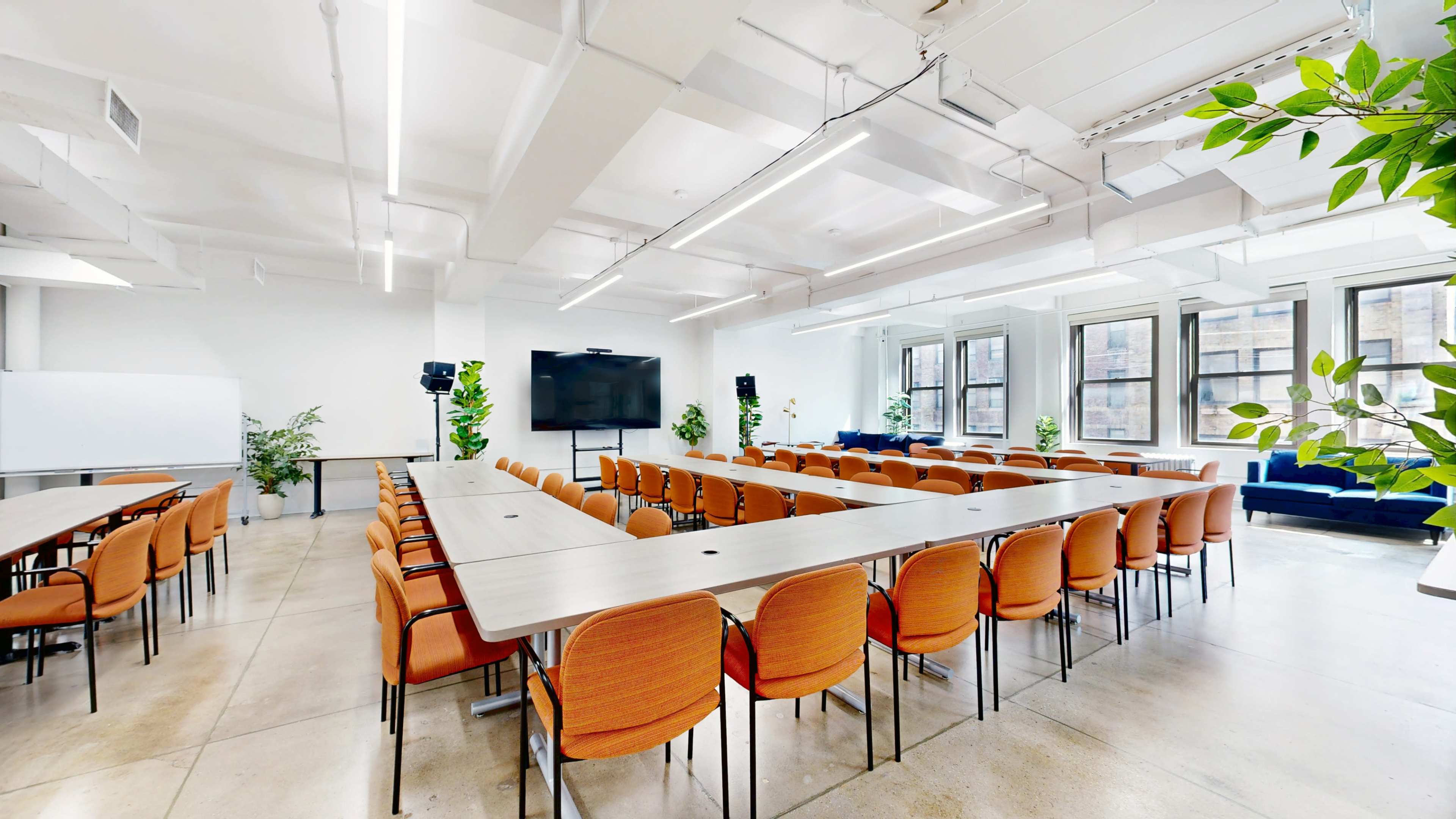 A well-lit conference room features several rows of orange chairs arranged around a long, rectangular table with a large screen and plants in the background.