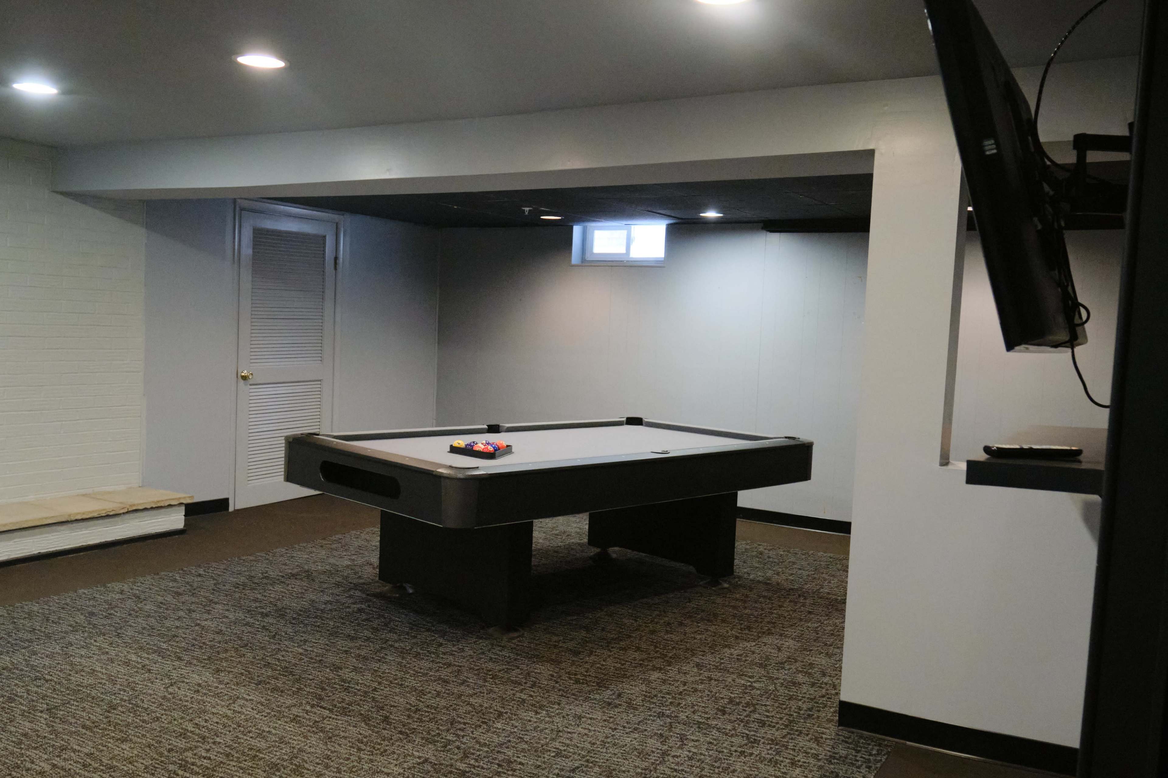 A spacious basement game room features a pool table in the center, with a television mounted on the wall and a small window letting in natural light.