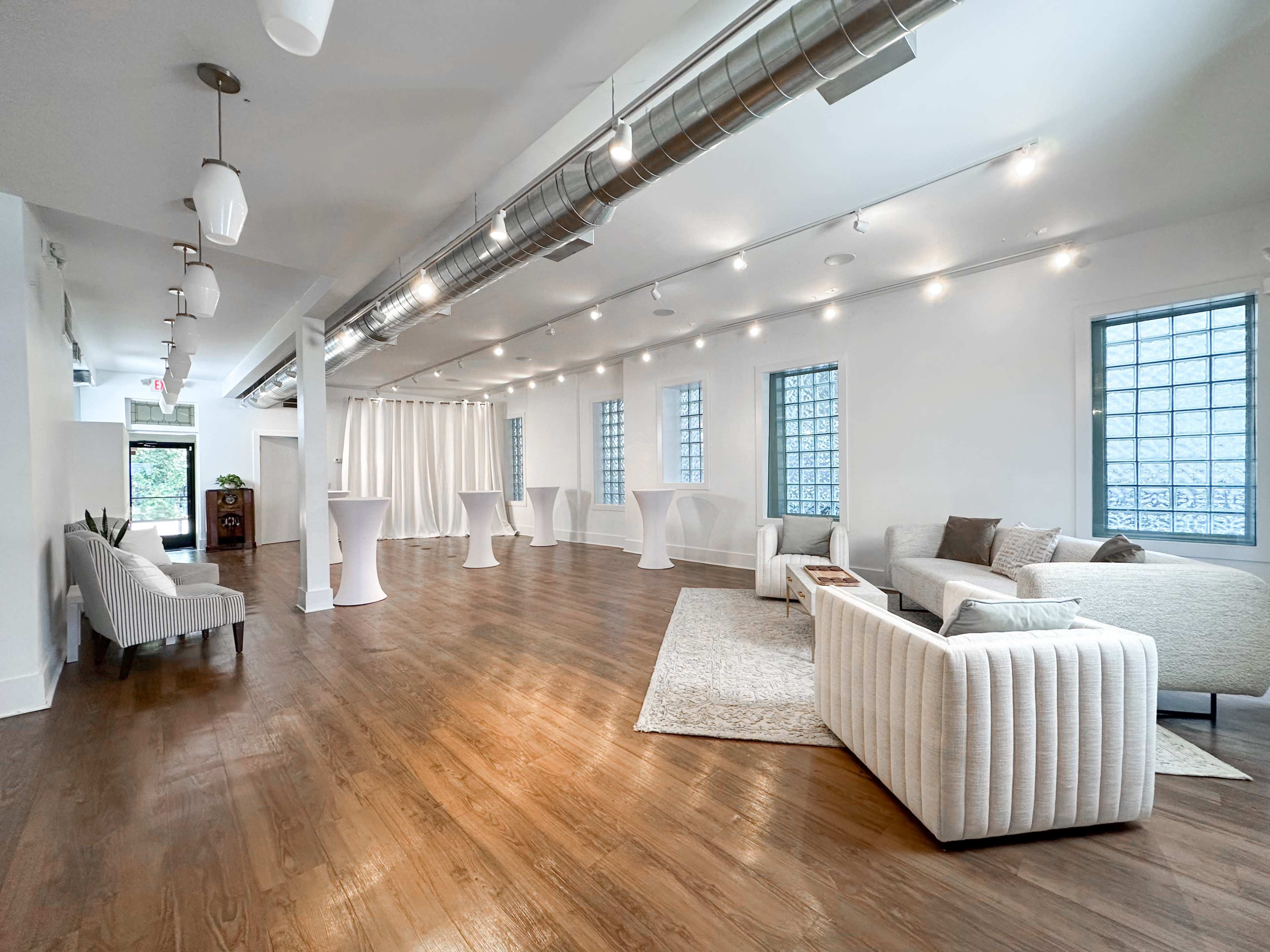 The interior of a spacious event venue features light wood flooring, white walls, and several seating areas set up with modern furniture and decor.