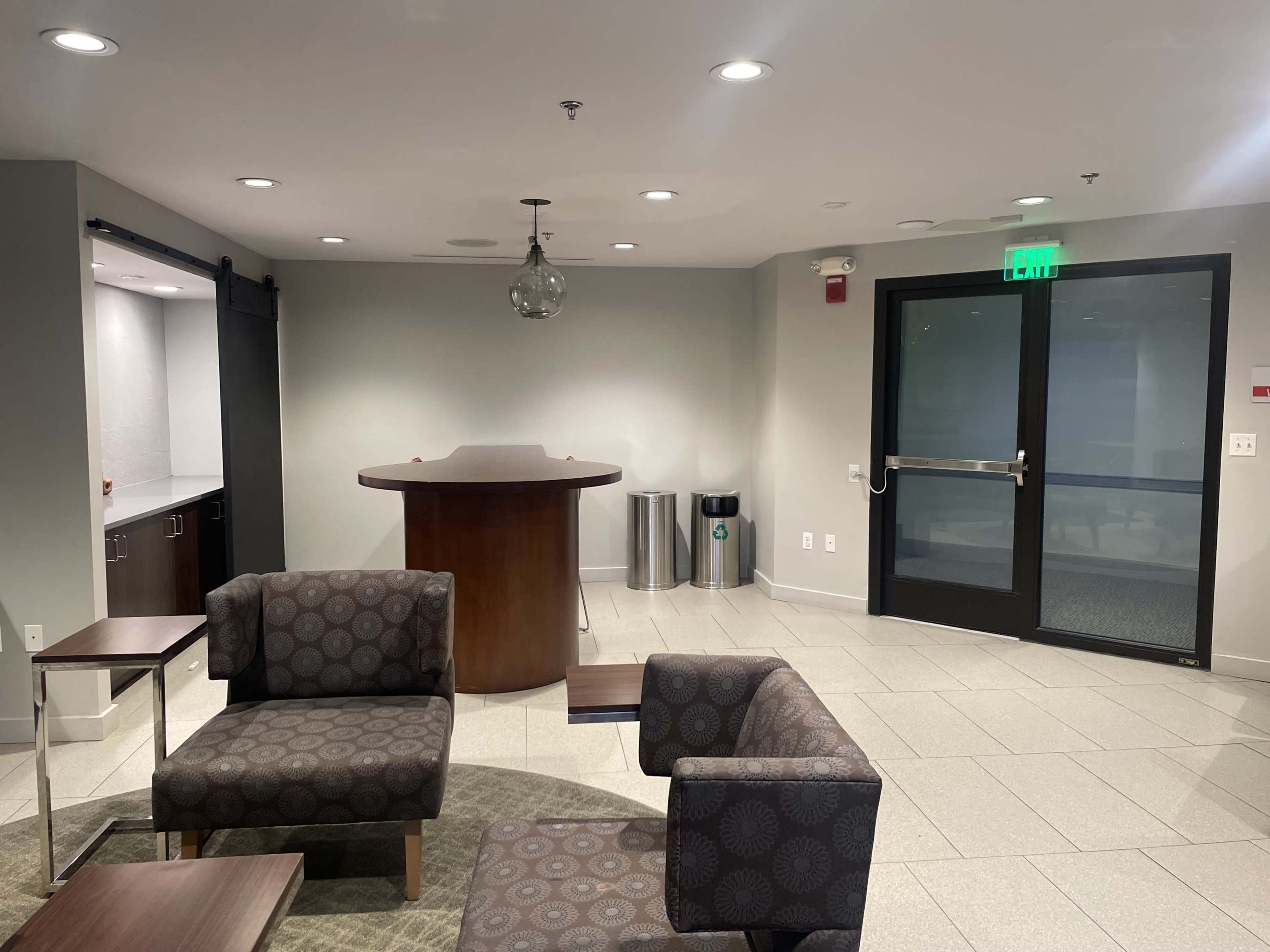 A modern lounge area with patterned chairs, a circular table, and a bar-like counter, next to a glass door and waste receptacles.