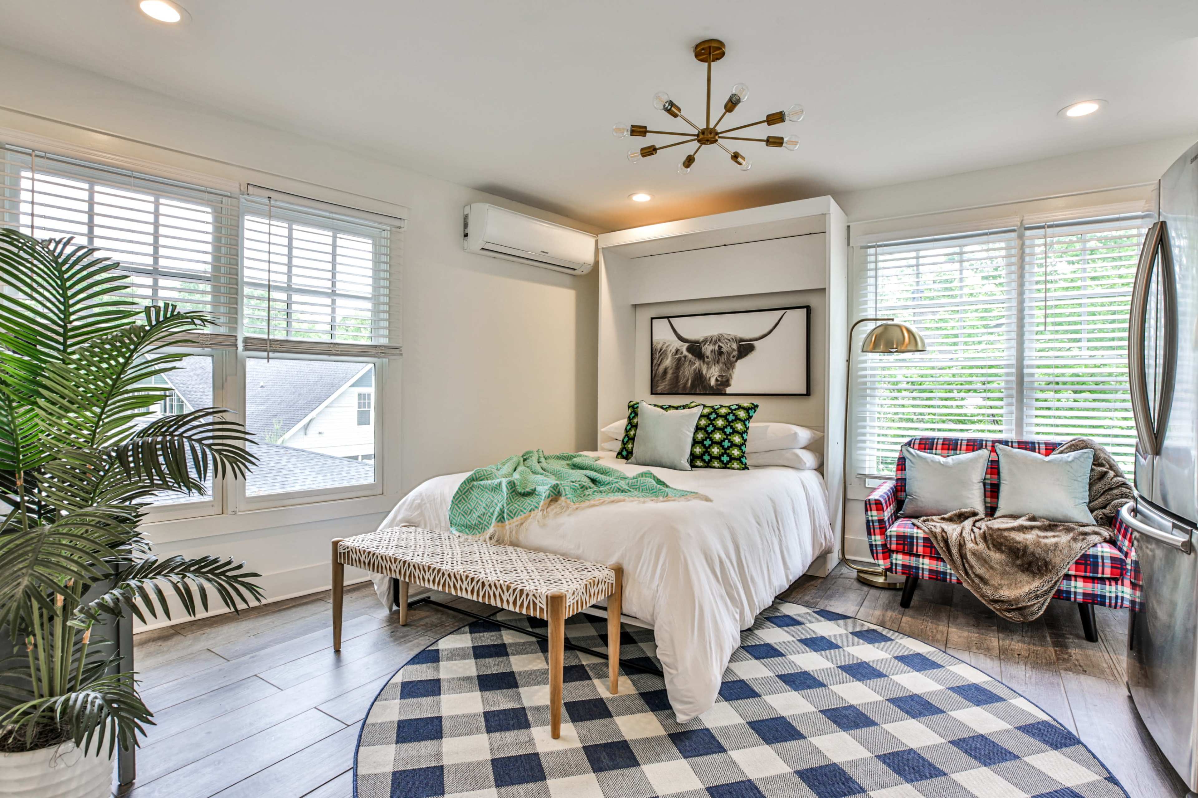 A cozy bedroom featuring a bed with green and white pillows, a decorative chair, and a large artwork on the wall, illuminated by natural light through window blinds.