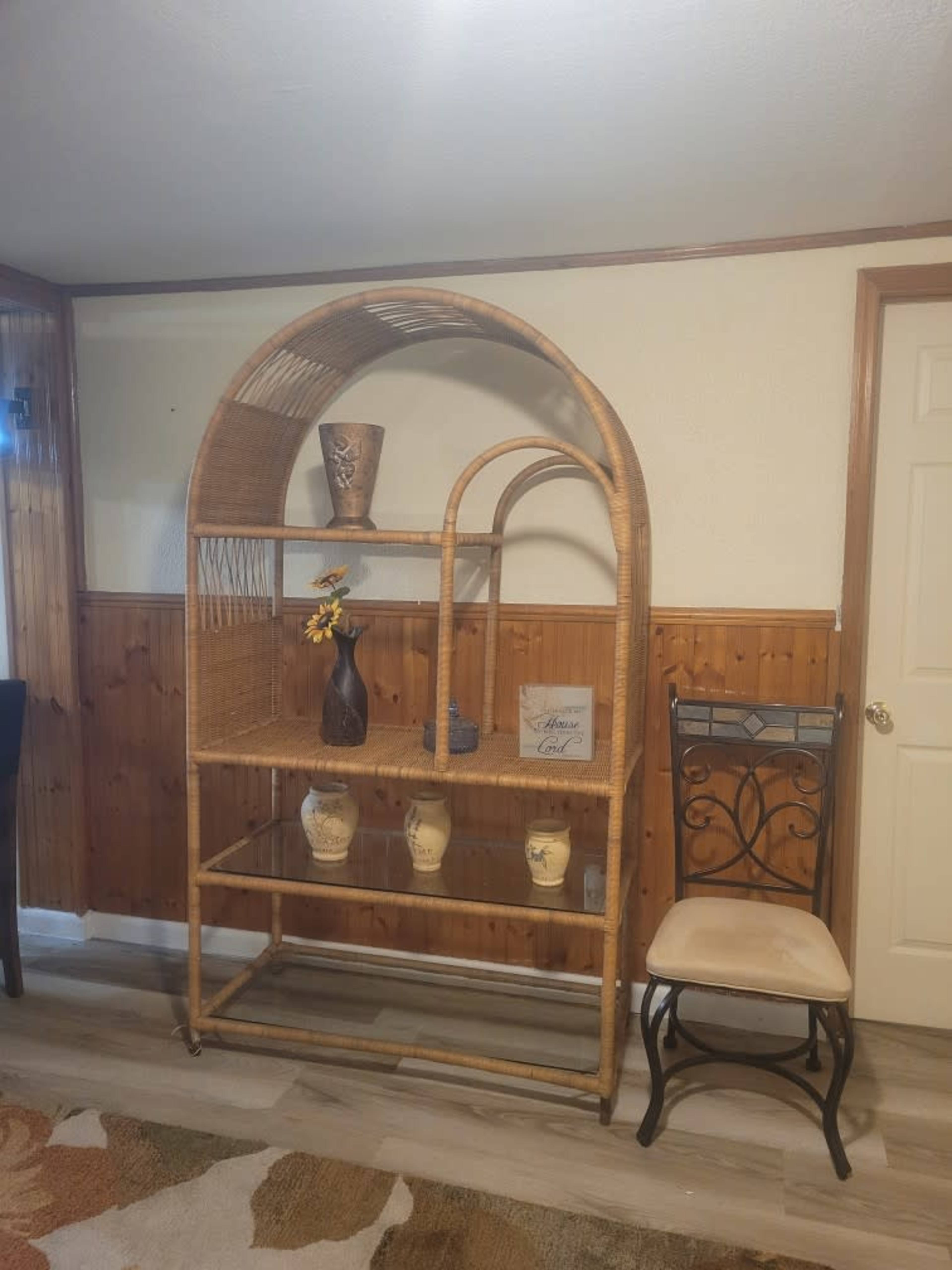 A rattan shelving unit stands against a wooden wall, featuring decorative vases and a chair nearby.