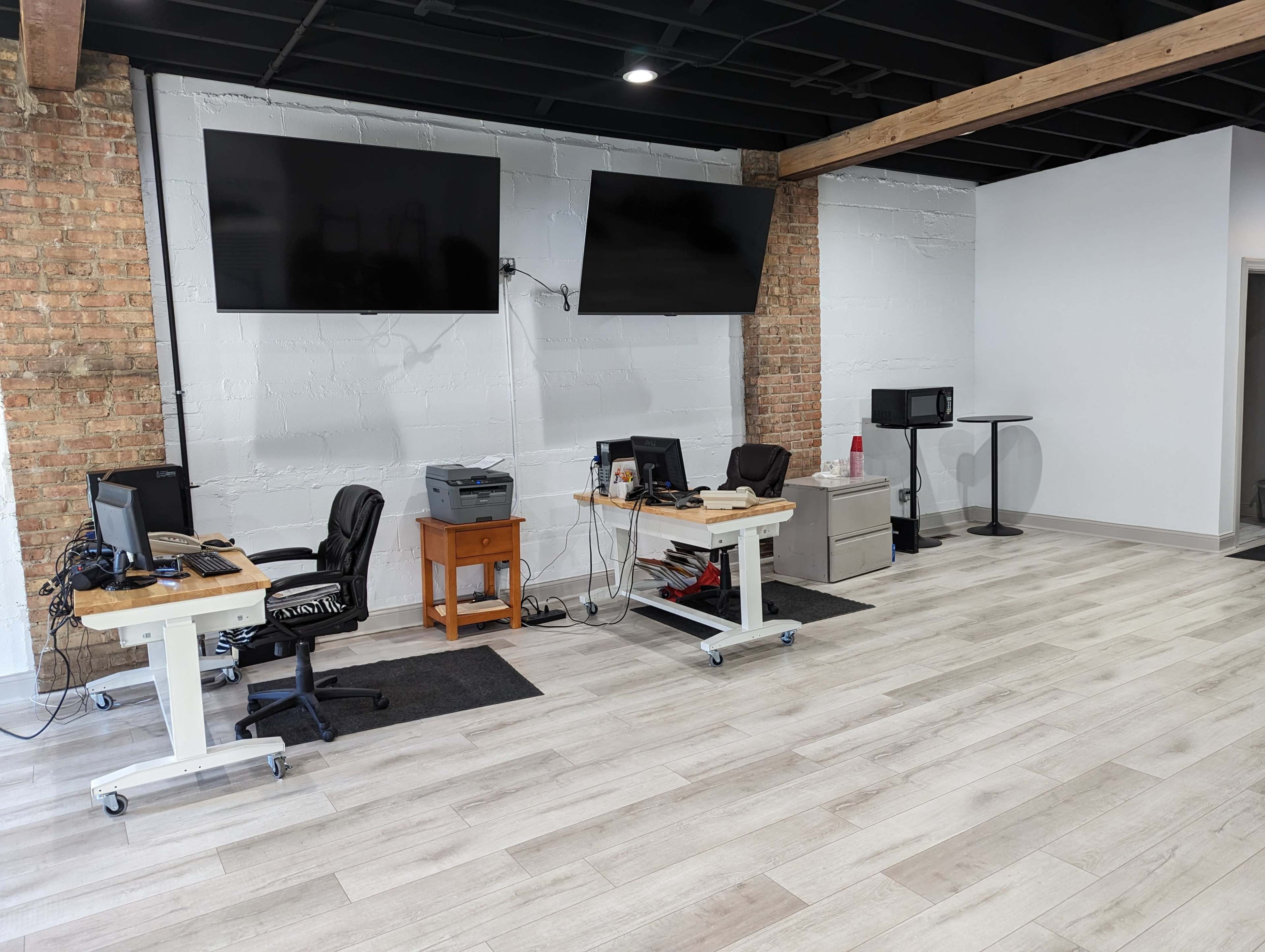 An office space with two desks equipped with computers, a filing cabinet, and two mounted television screens on the wall, all set against a backdrop of exposed brick and light-colored flooring.