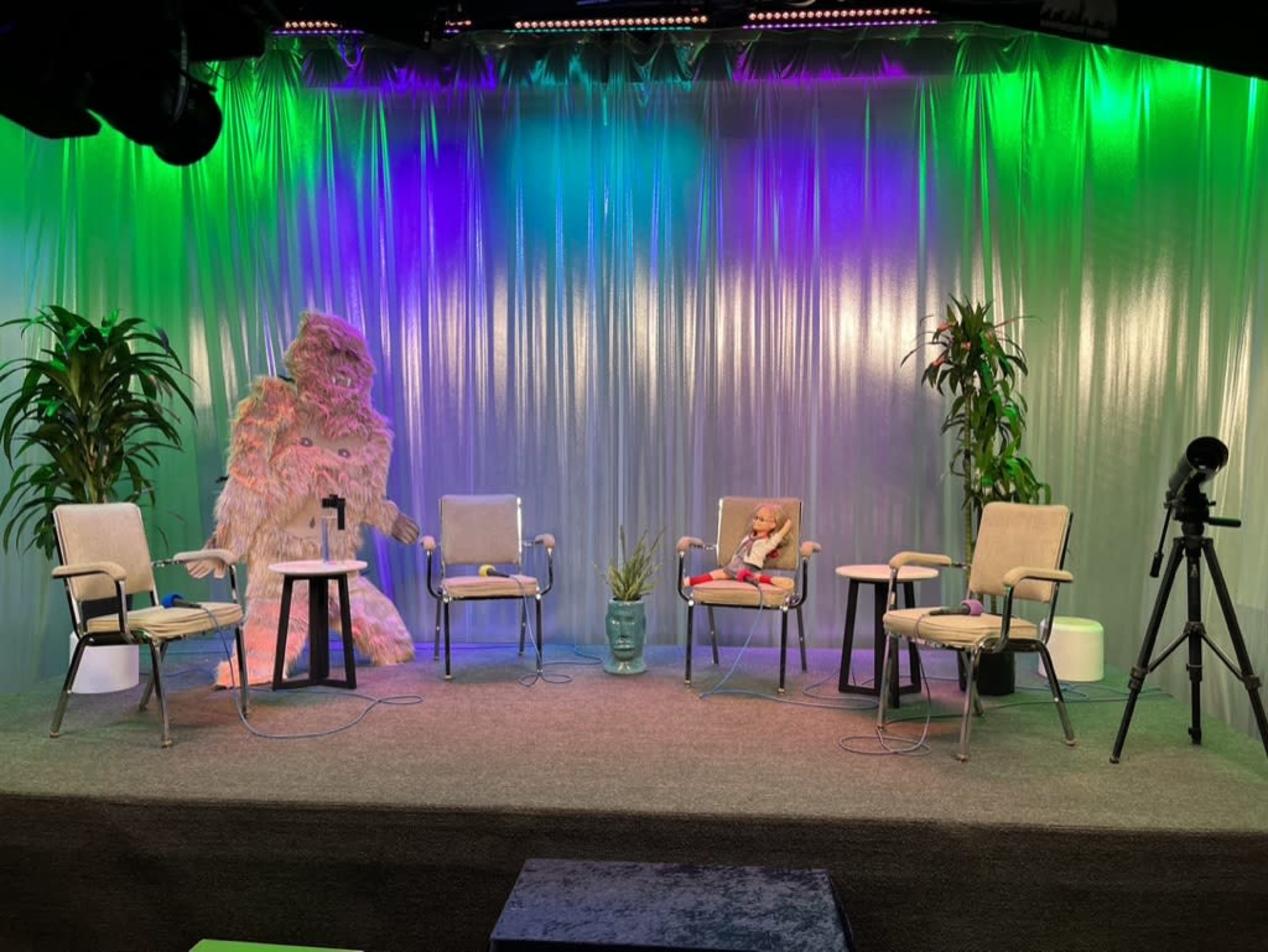 The scene features an empty stage with several chairs arranged in front of a colorful backdrop, accompanied by a large decorative figure and a camera on a tripod.